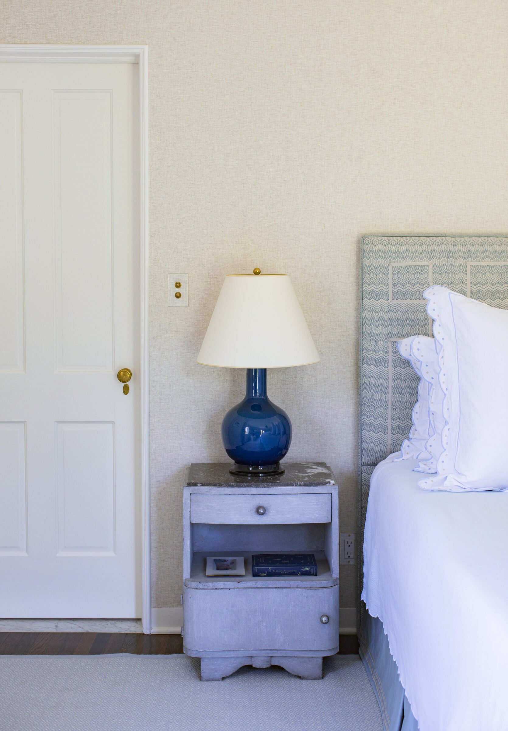 Guest bedroom with a blue upholstered headboard, a blue lamp on a gray painted bedside chest and a white paneled door