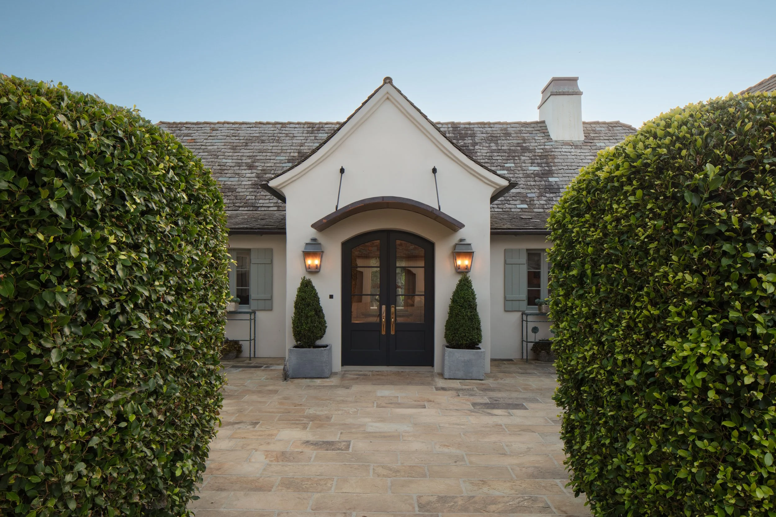 White house front with an arched gabled entry, dark double doors, clipped hedges and a stone courtyard