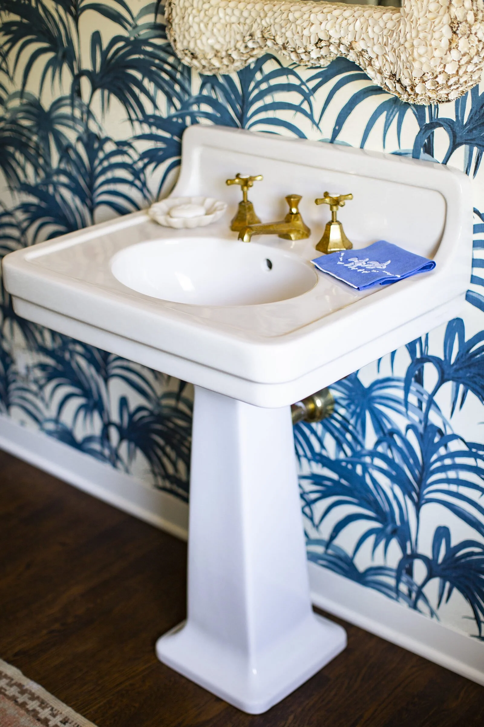 Pedestal sink with brass faucets and a blue embroidered towel against blue palm-frond wallpaper