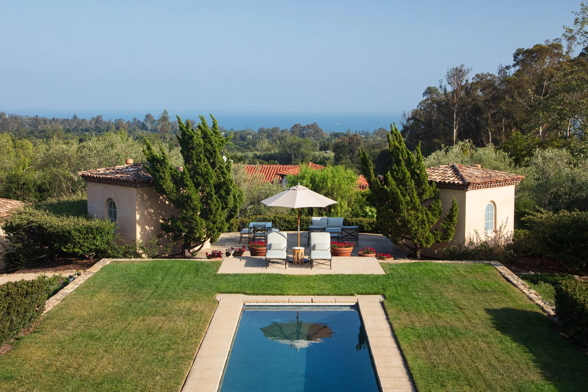 Garden pool with a patio umbrella and lounge chairs, framed by cypress trees with a glimpse of the ocean beyond