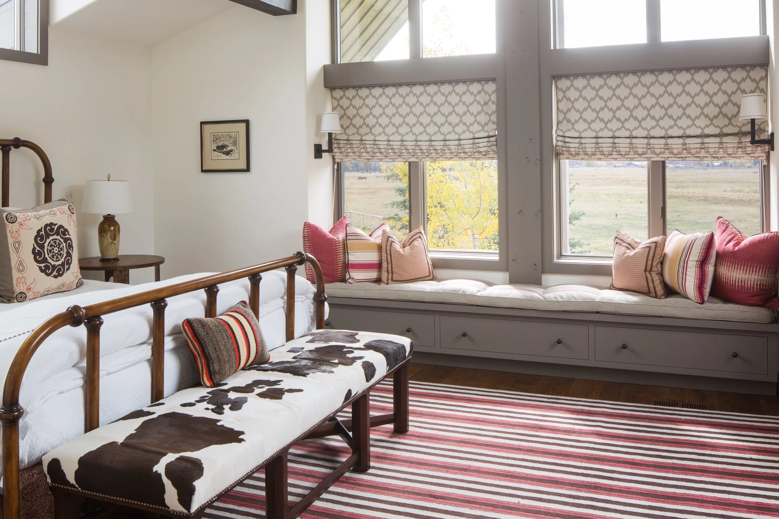 Window seat with a gray cushion, a cowhide bench, a striped rug and patterned Roman shades looking out to a meadow