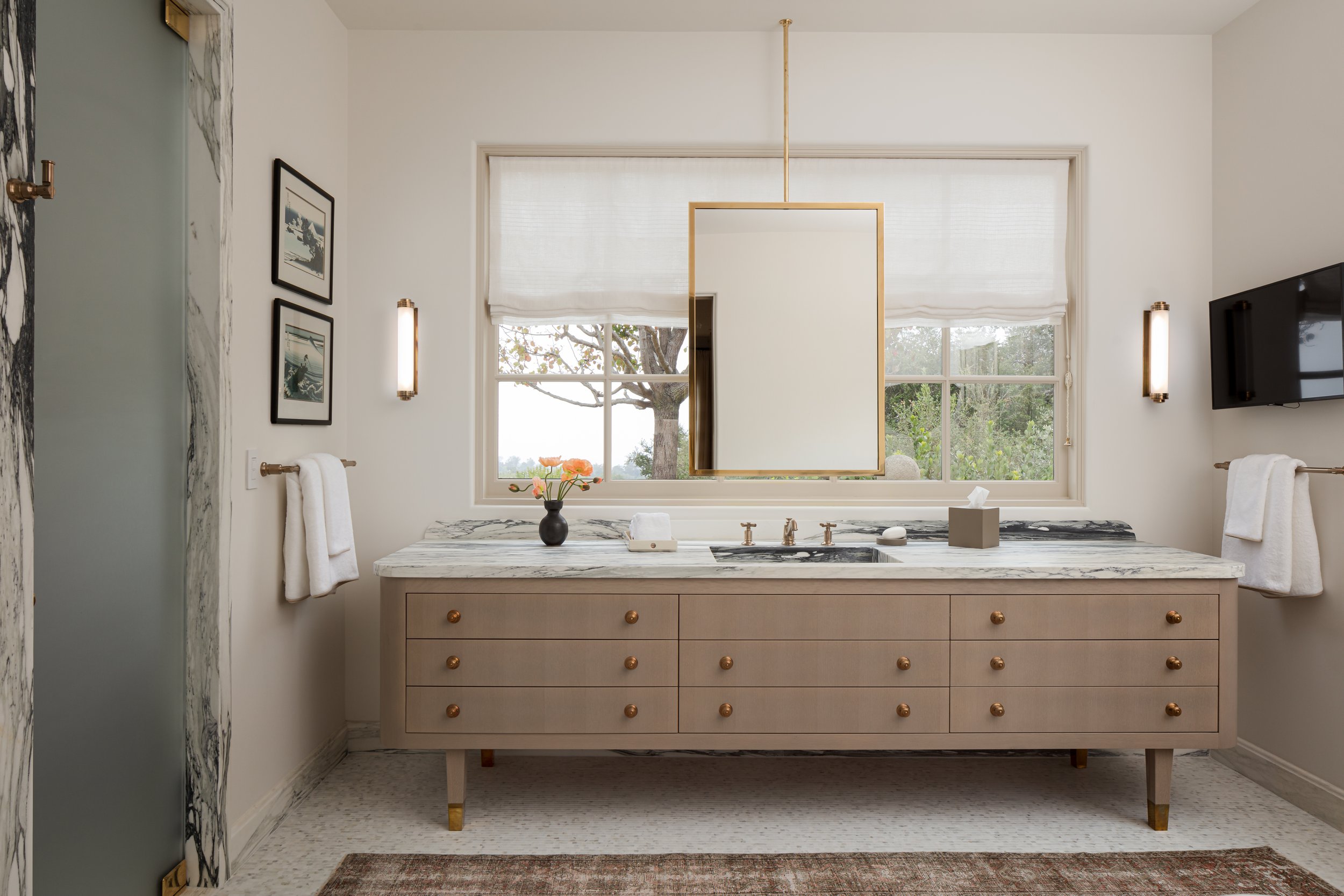 Primary bath with a long taupe double vanity, a brass-framed mirror, wall sconces and a window with a white shade