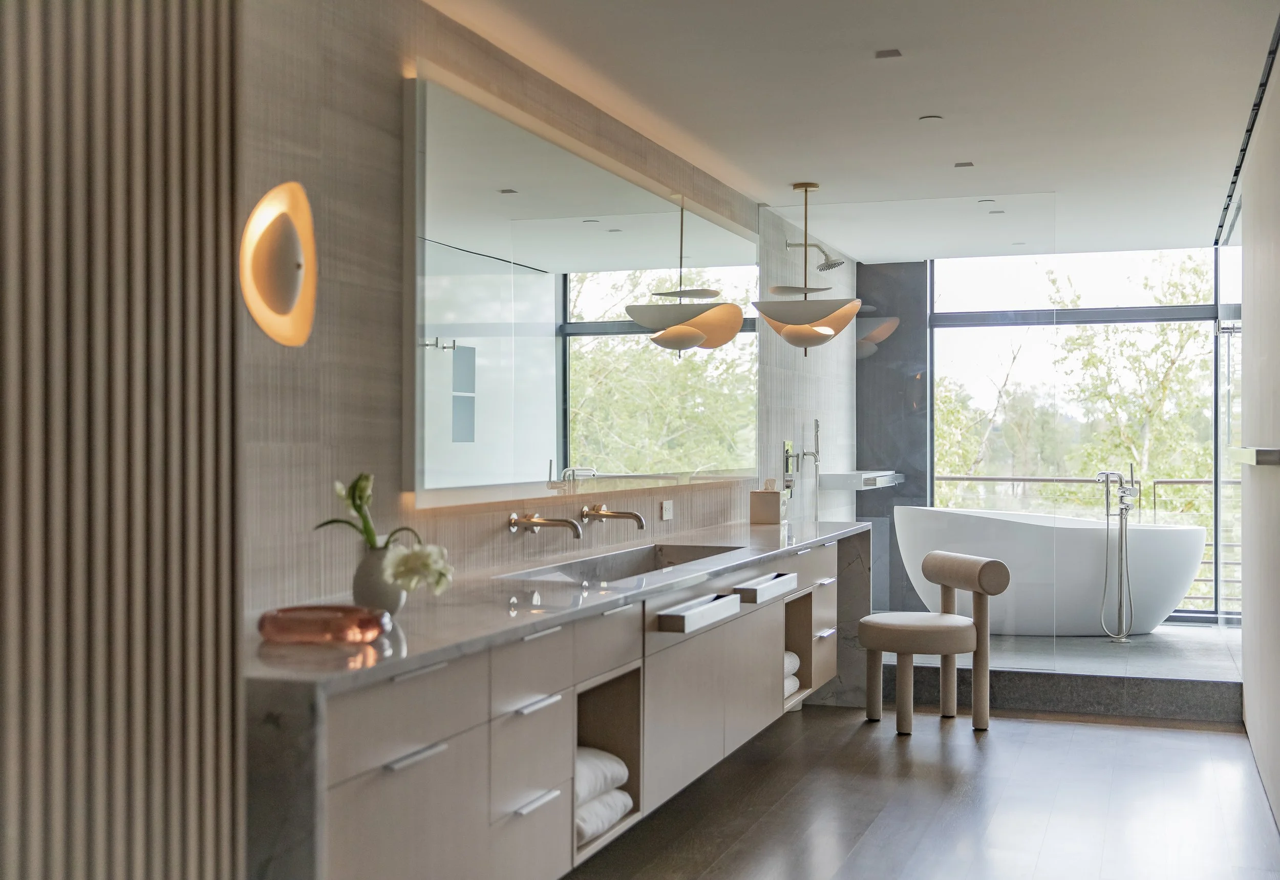 Primary bath with a long oak vanity, glowing mirror and freestanding tub by a window
