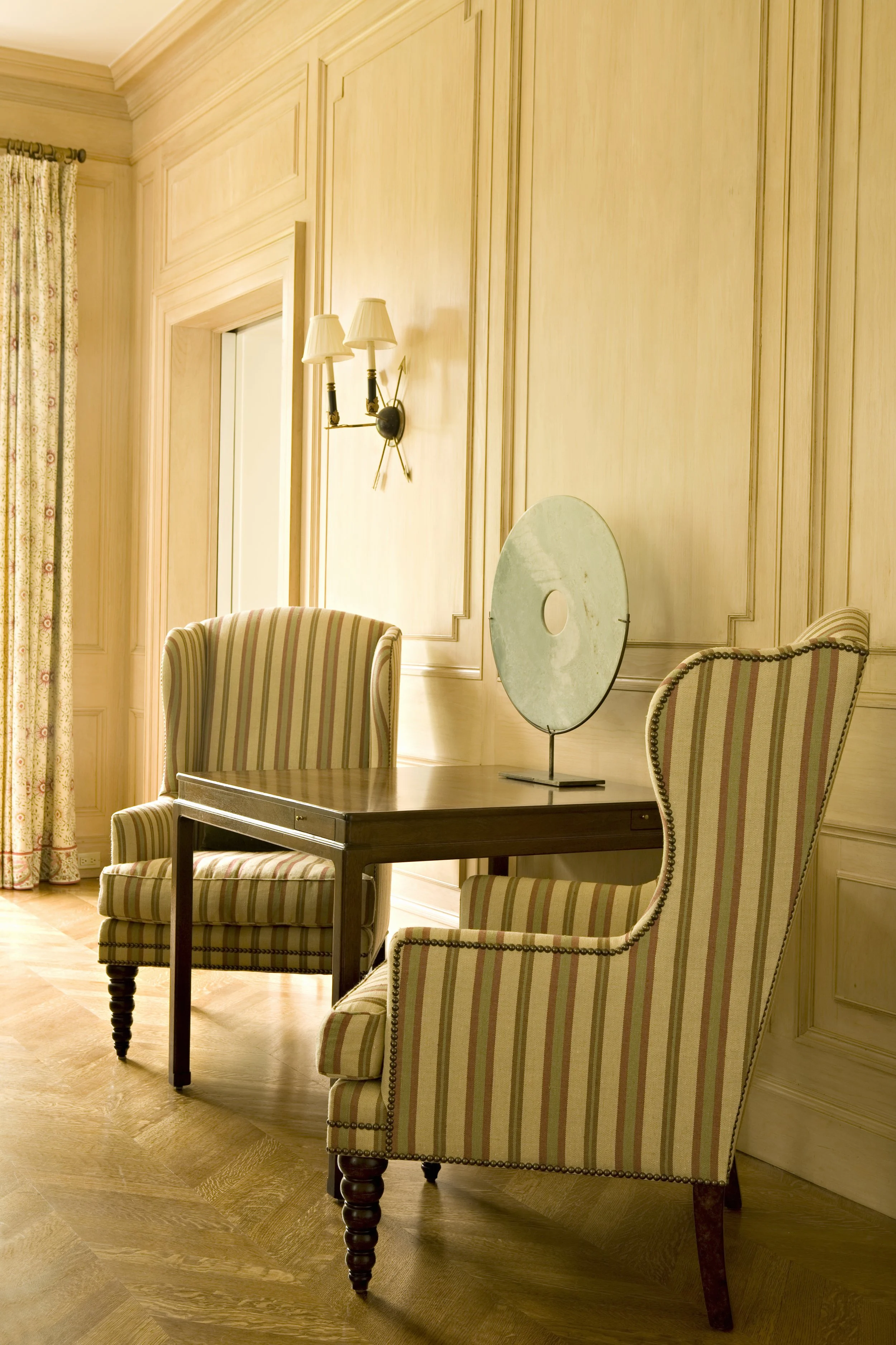 Striped wing chairs and a desk with a sculpture in a warm paneled room