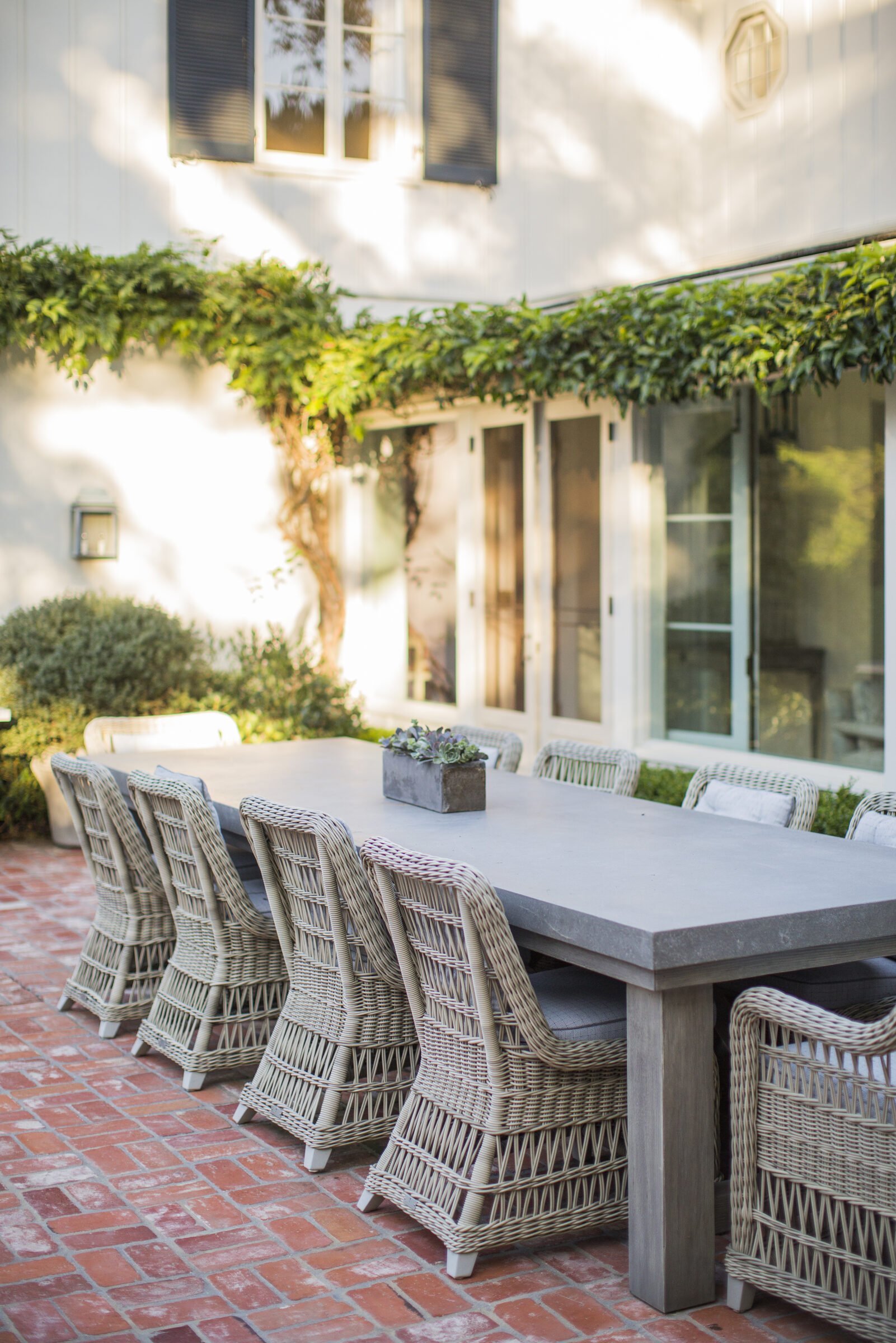 Outdoor dining table with woven wicker chairs on a herringbone brick patio under a canopy of climbing vines