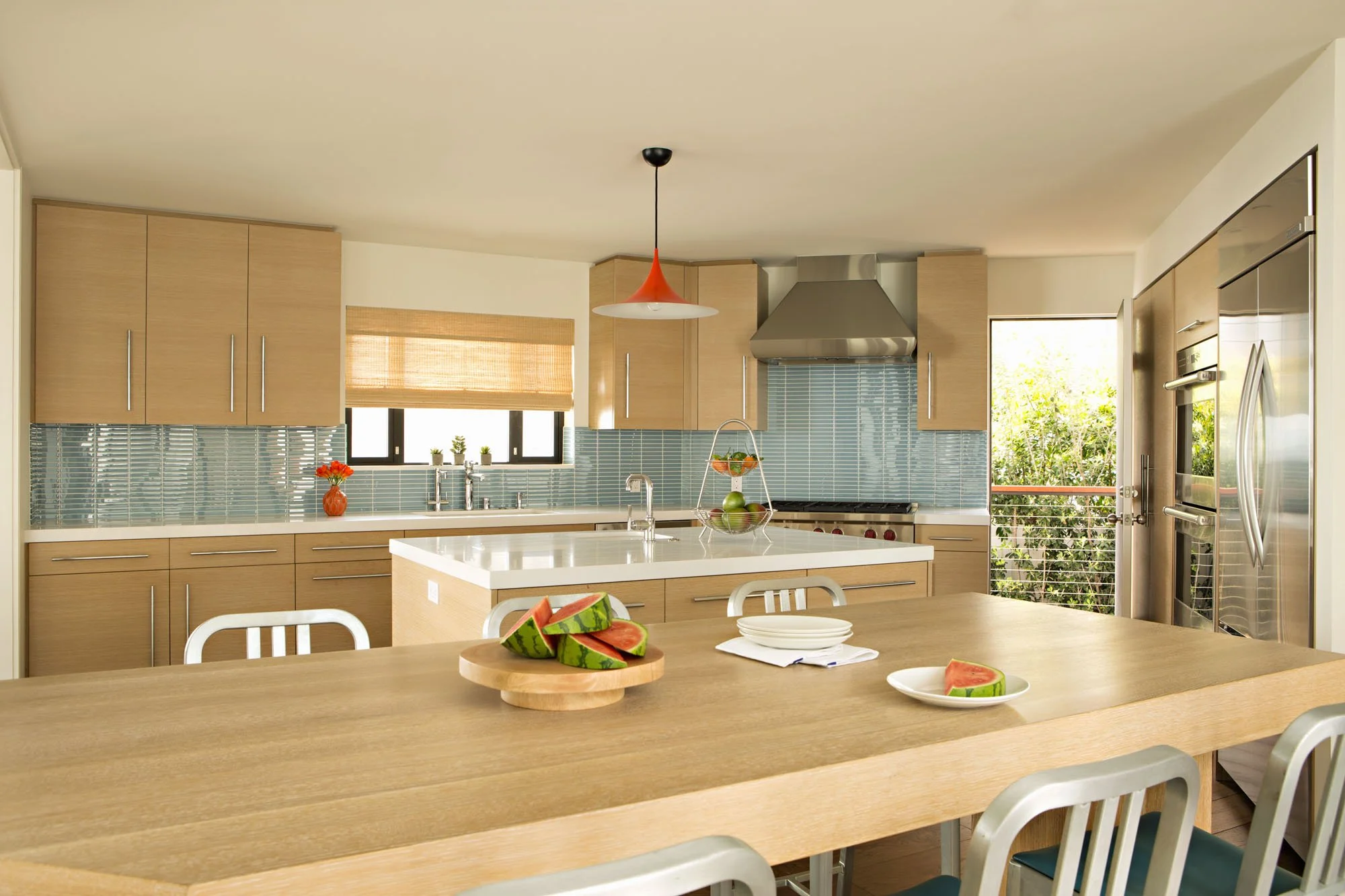 Kitchen with light oak cabinetry, pale blue glass tile, a red pendant light and aluminum stools at a long island