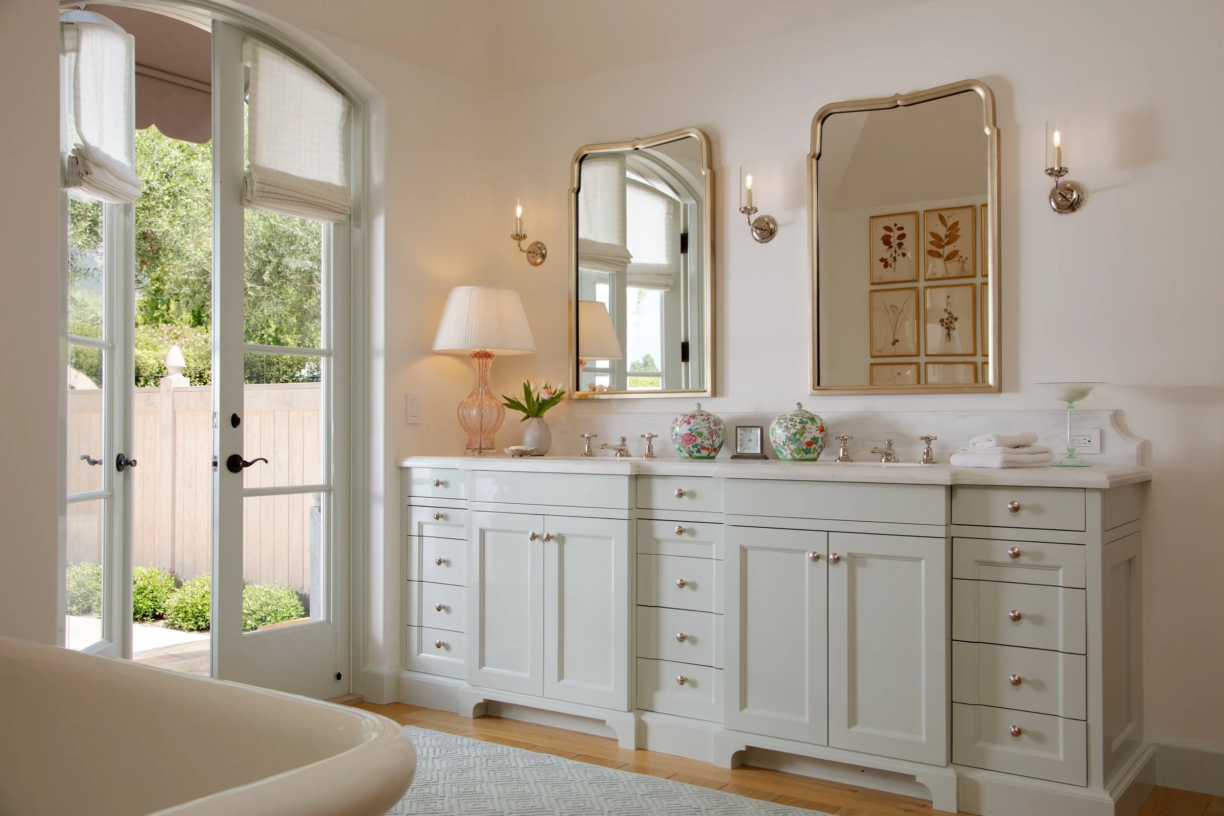 Bath vanity with pale green cabinetry, two gilt mirrors framing pressed botanical prints and a French door to the garden