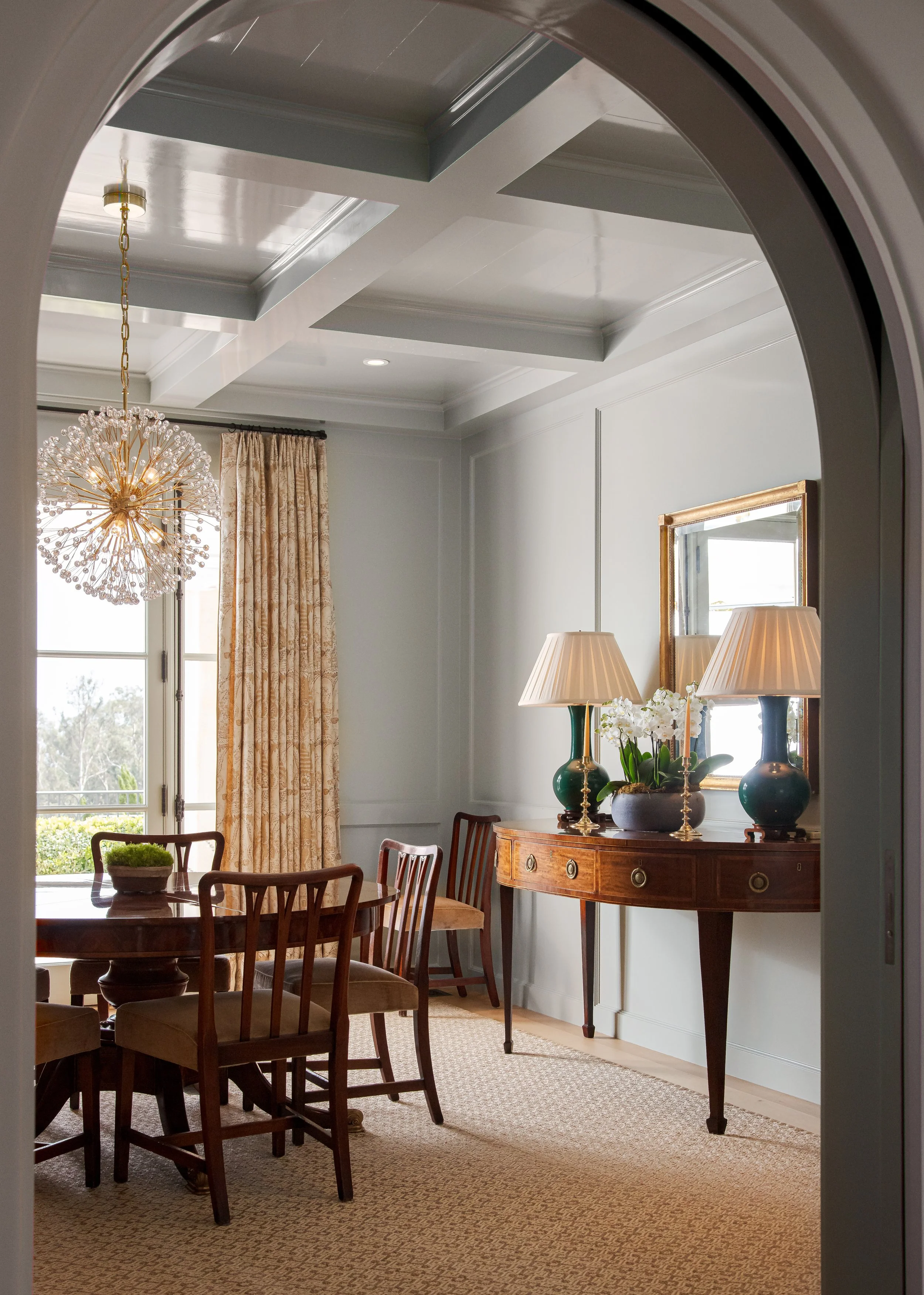 Dining nook with a coffered ceiling, a crystal chandelier, wood chairs and a console with two lamps beneath a gilt mirror