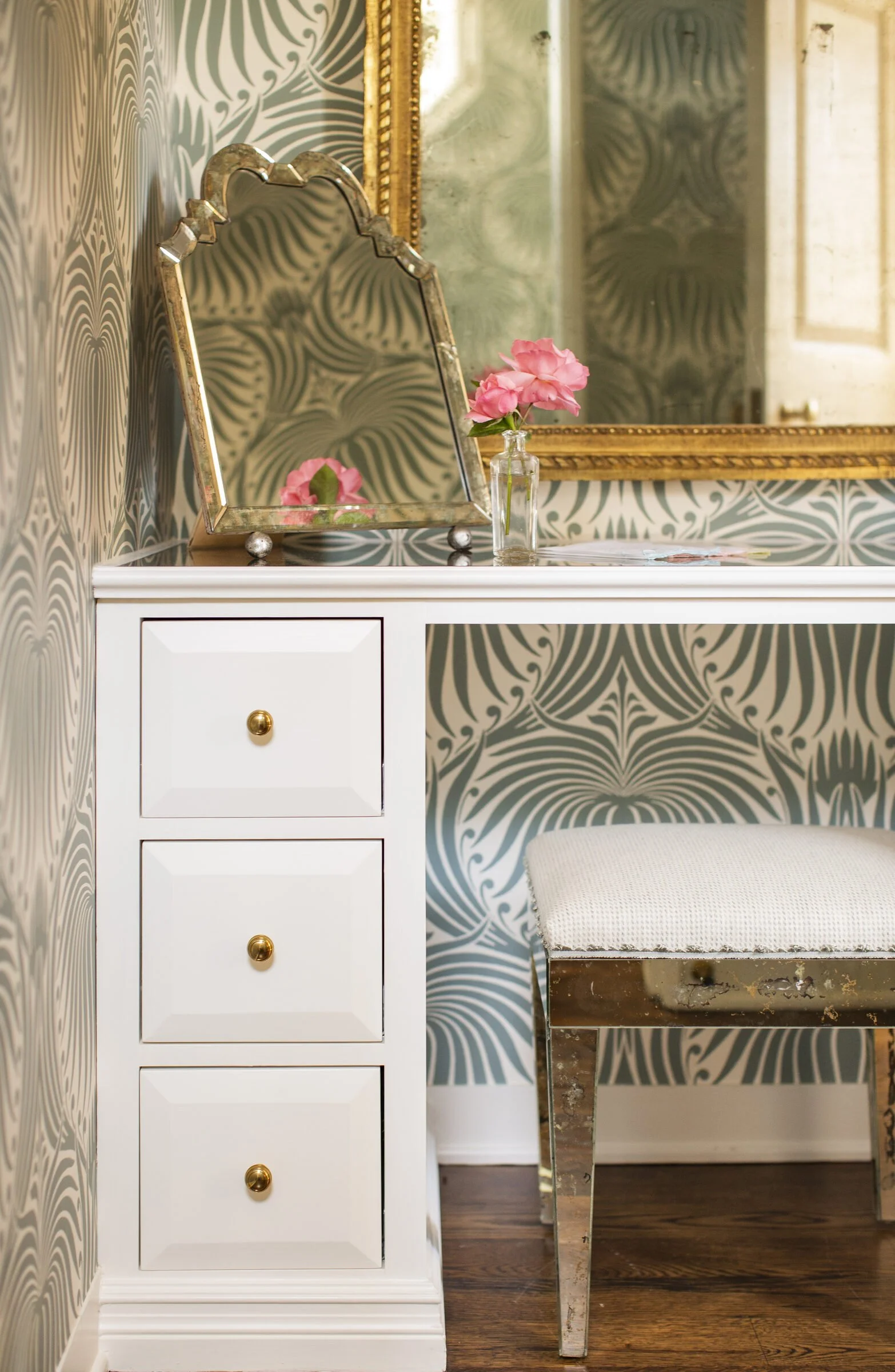 Dressing nook with a white three-drawer vanity, a mirrored bench, a gilt mirror and green-gray patterned wallpaper