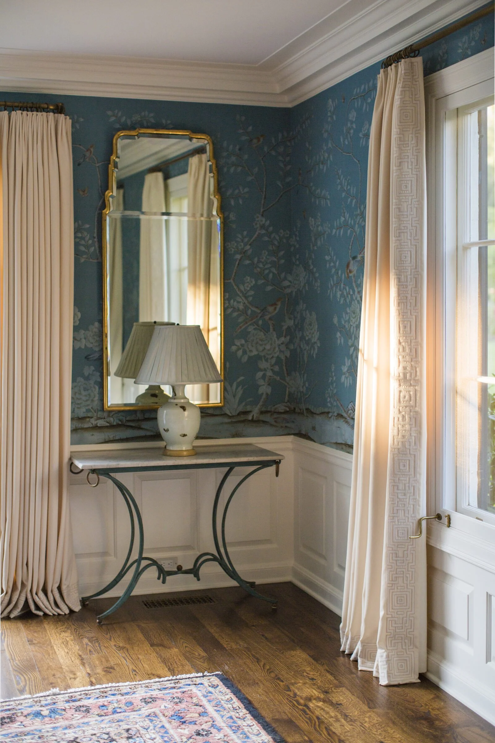 Sitting room with blue chinoiserie wallpaper, long cream curtains, a gilt mirror, a console table and an antique rug