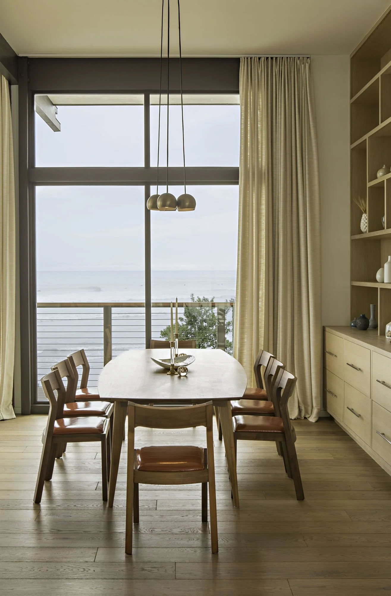 Dining table with a pendant cluster and wood chairs beside floor-to-ceiling windows facing the ocean