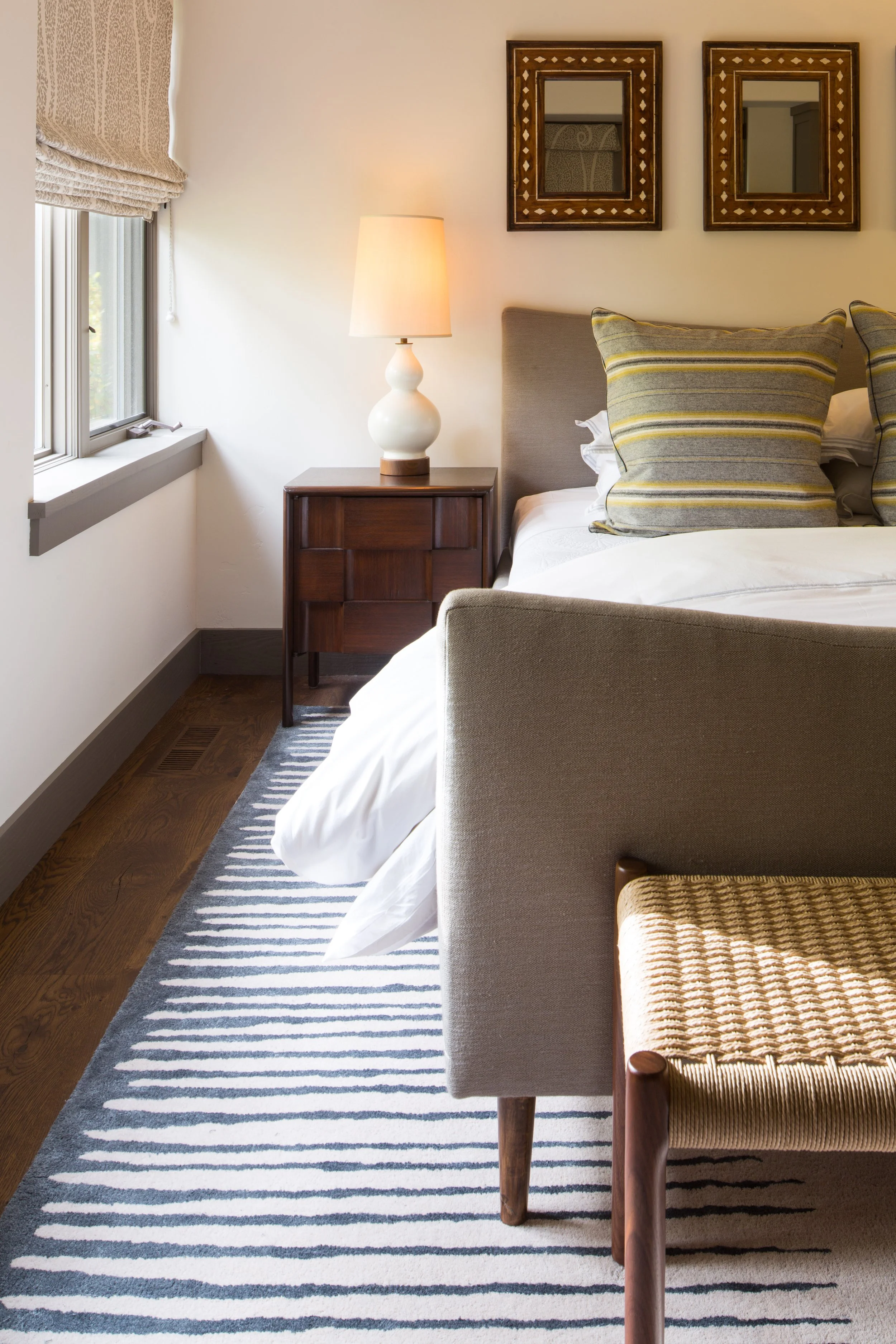 Bedroom corner with an upholstered gray headboard, a striped pillow, a dark wood nightstand and a striped runner