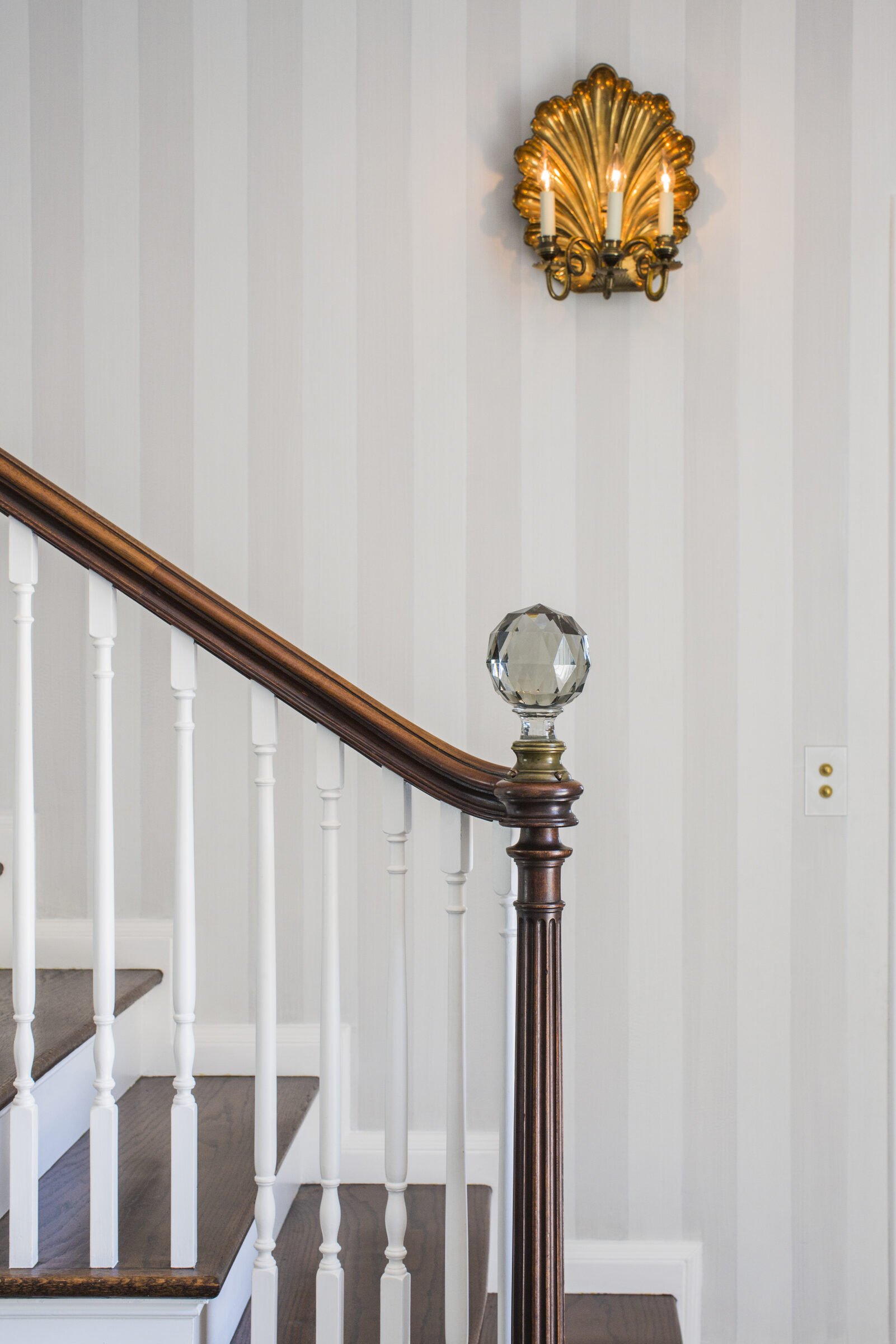 Staircase with a wood handrail, white balusters, a faceted crystal newel post and a gilded shell sconce against striped wallpaper