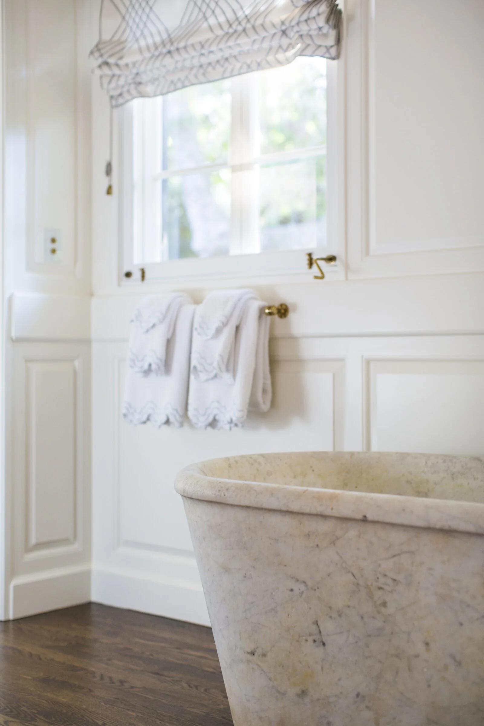 Freestanding stone tub beside white paneled walls and a window with a patterned Roman shade