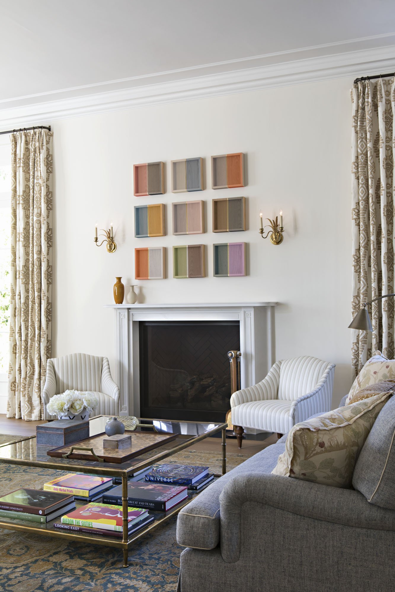 Living room with a gray painted fireplace, striped armchairs and a grid of colorful art above the mantel