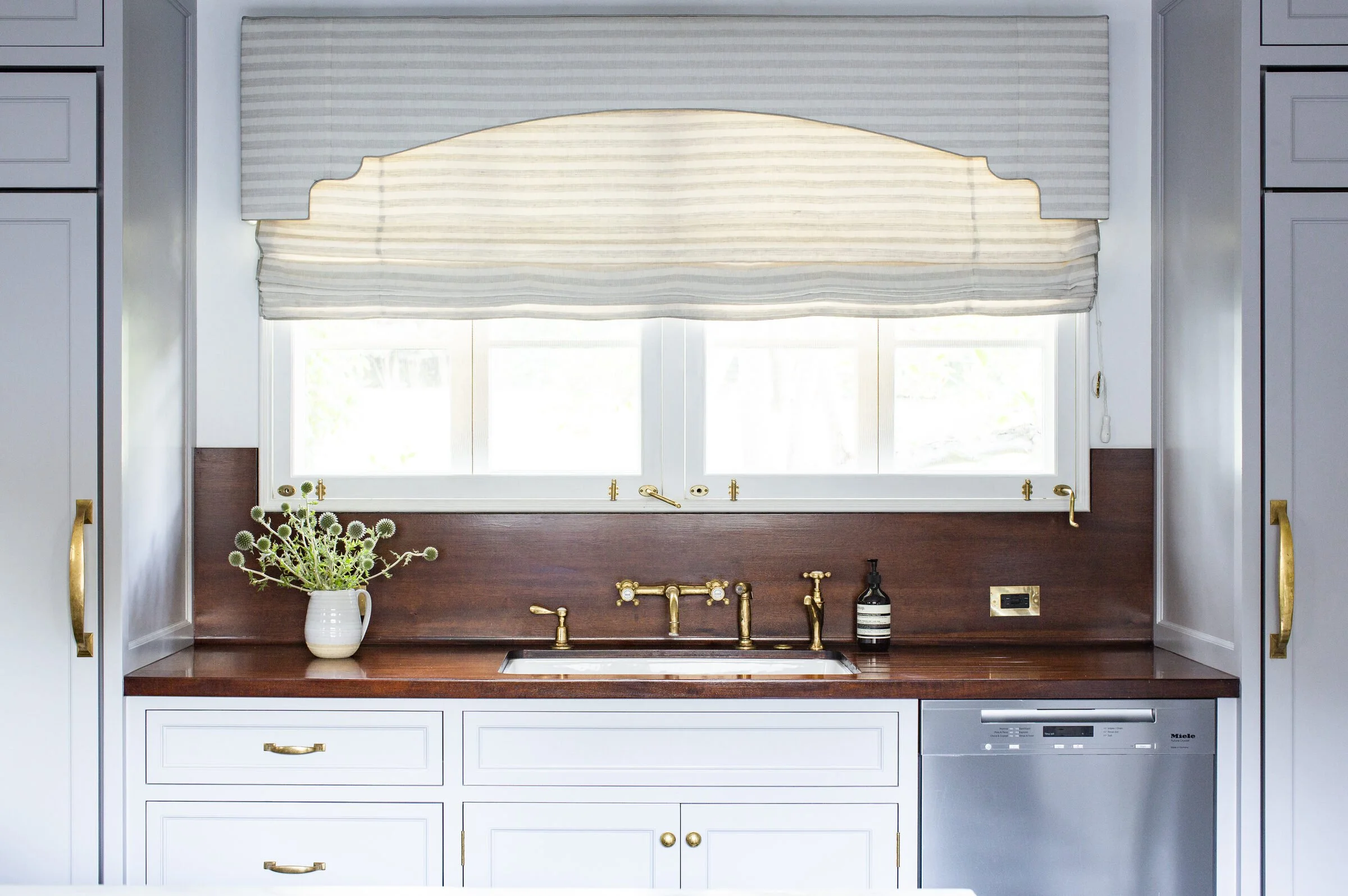 Kitchen window with a scalloped striped shade, a dark wood backsplash, unlacquered brass fixtures and blue-gray cabinetry