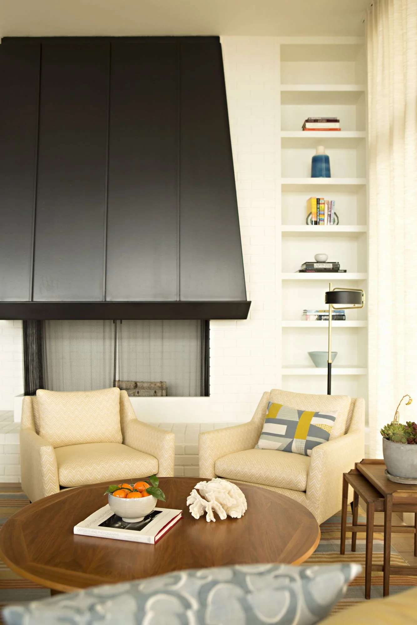 Living room with a dark metal fireplace hood, built-in shelves, two cream armchairs and a yellow and gray pillow