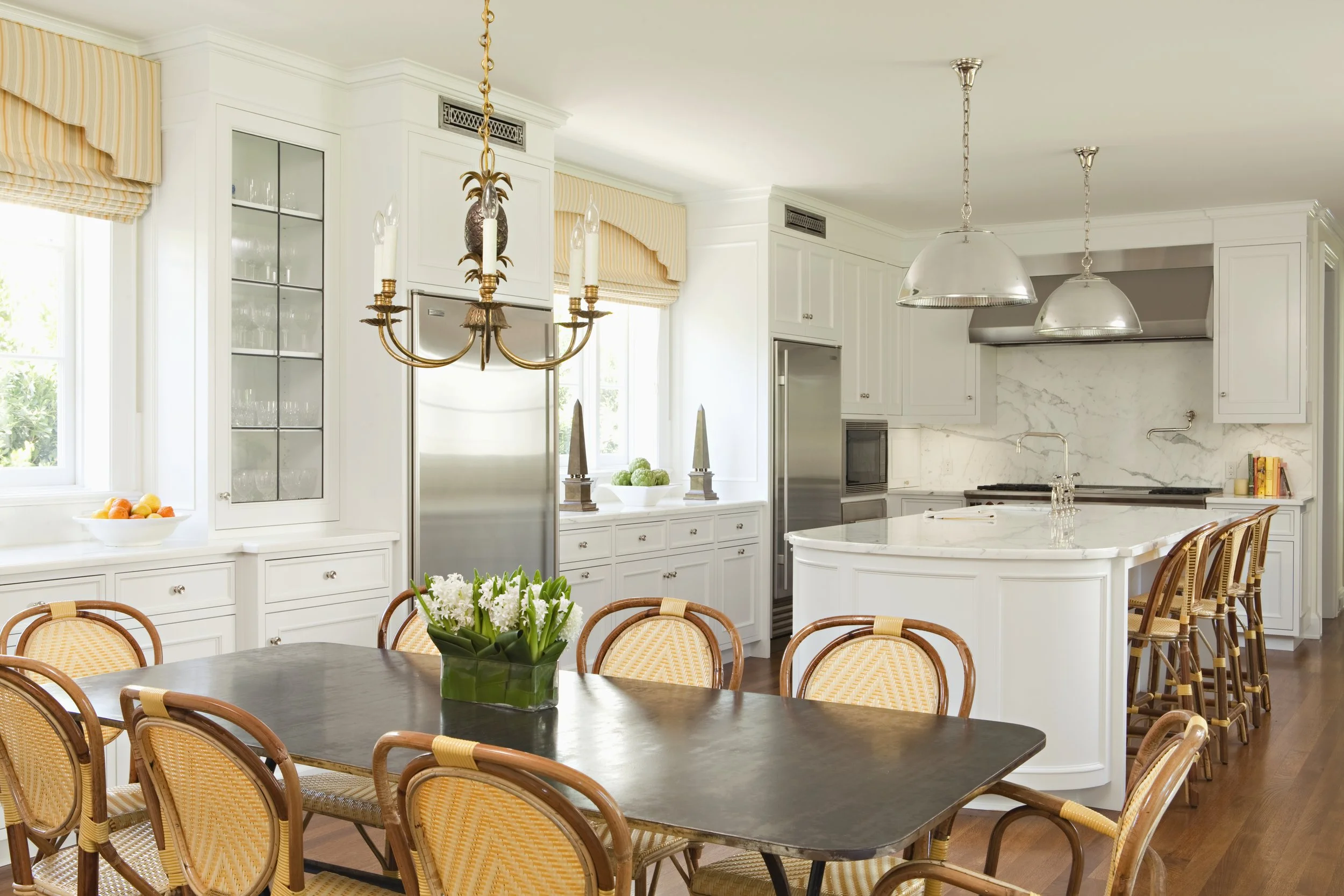 Kitchen with a white island, rattan dining chairs, brass chandelier and a dark dining table