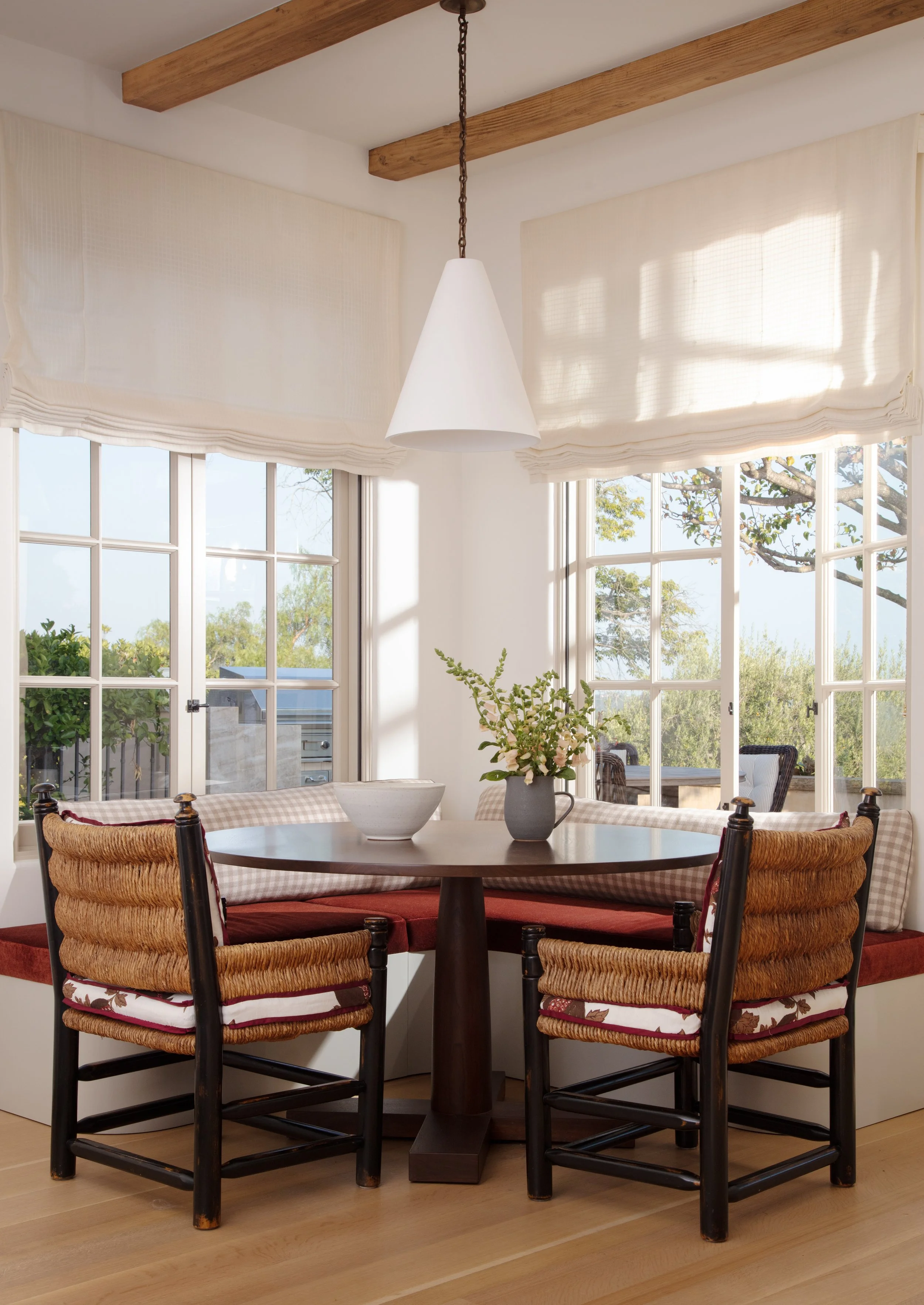Breakfast nook with a round table, rush-seat chairs, a red cushioned banquette and corner windows with garden views