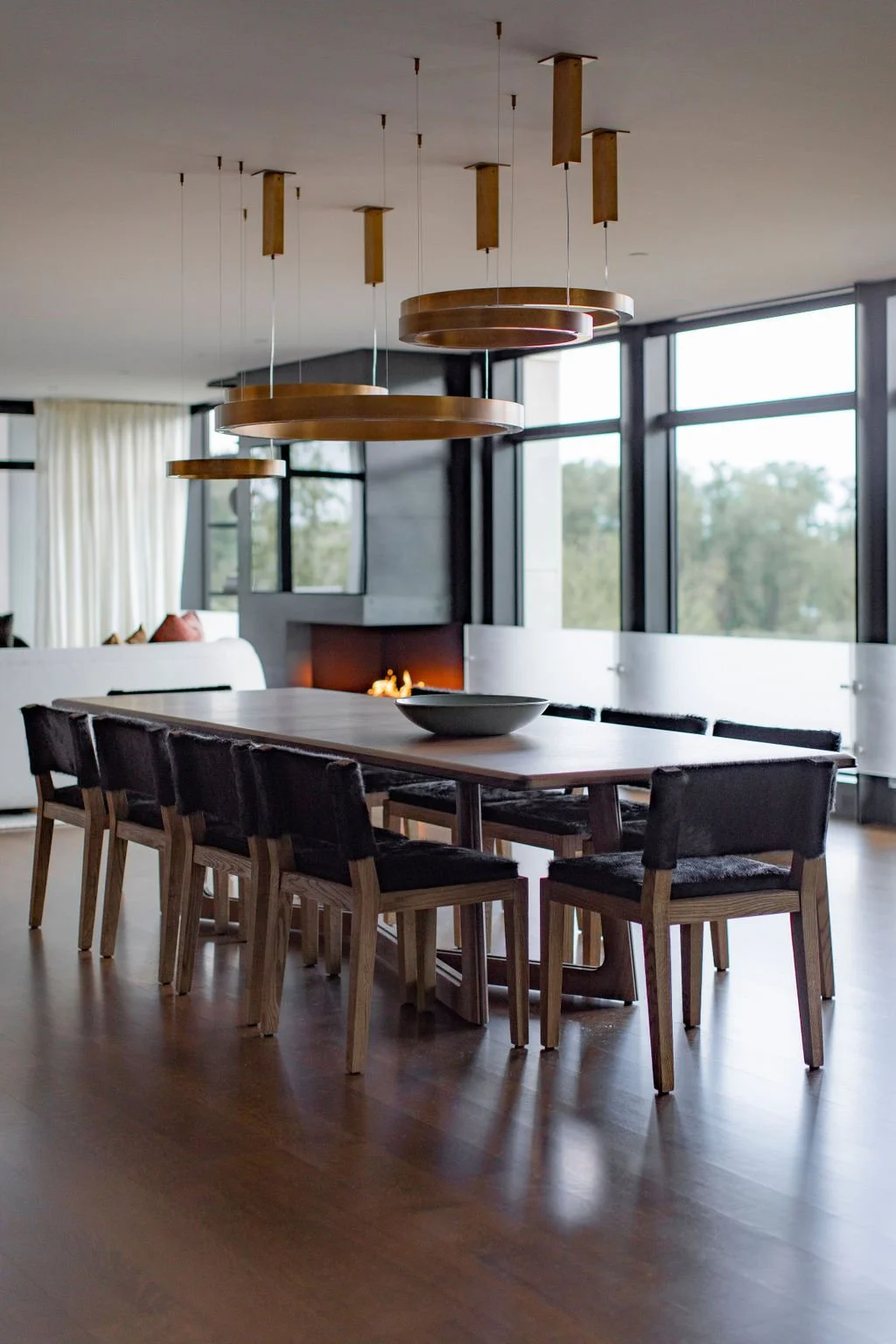 Dining room with a long wood table, dark leather chairs, copper pendants and a fireplace beside floor to ceiling windows