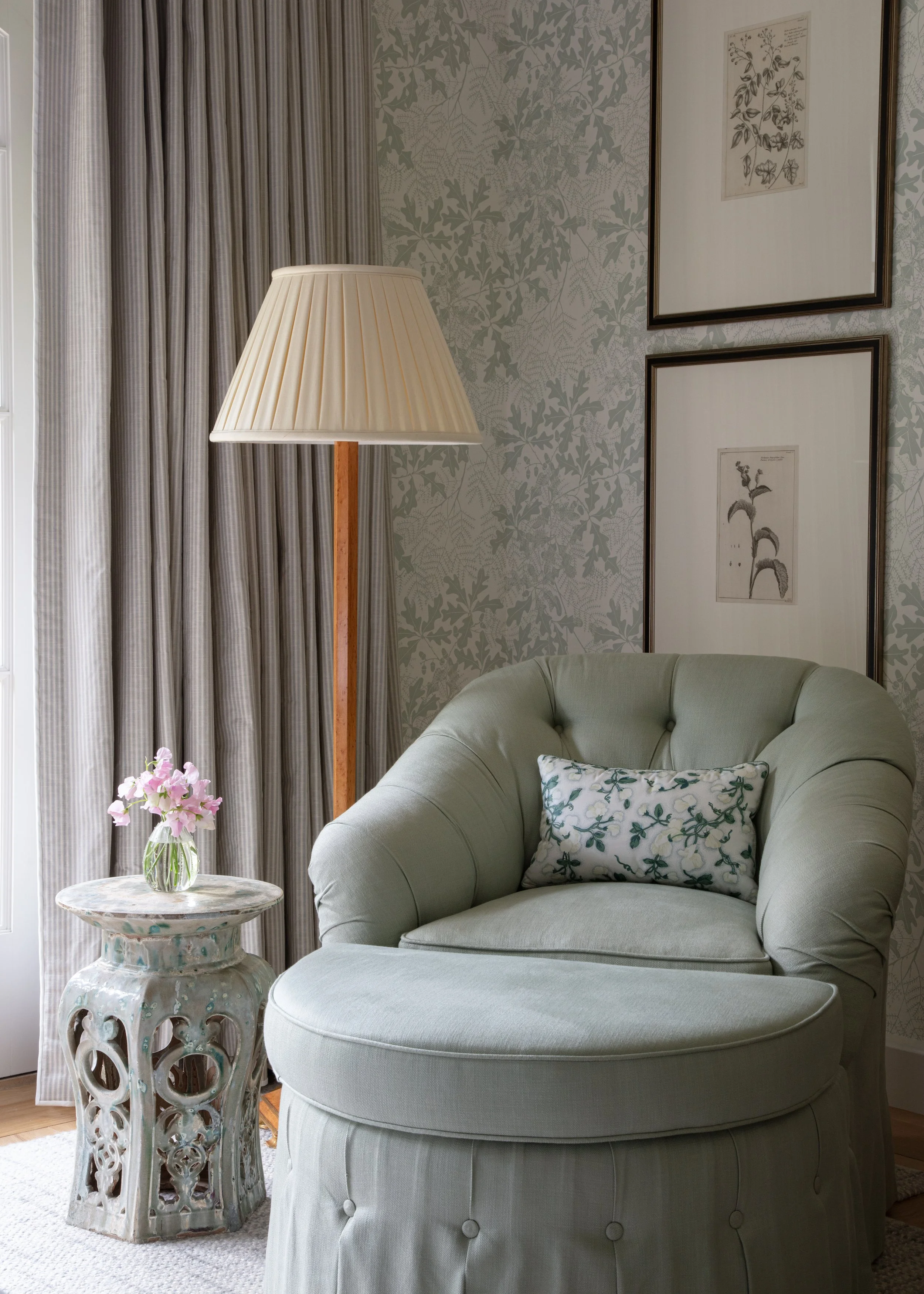 Sage green tufted armchair with a floral pillow, a pleated floor lamp and gray draperies against patterned wallpaper