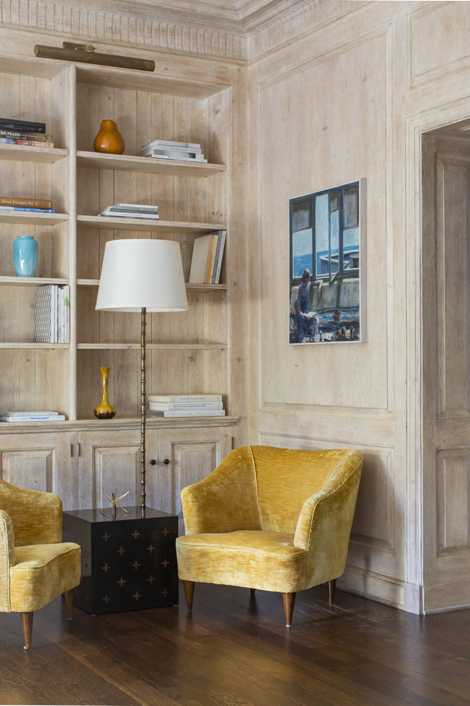 Built-in bookshelves in limed wood with a floor lamp, a framed seascape and mustard velvet armchairs