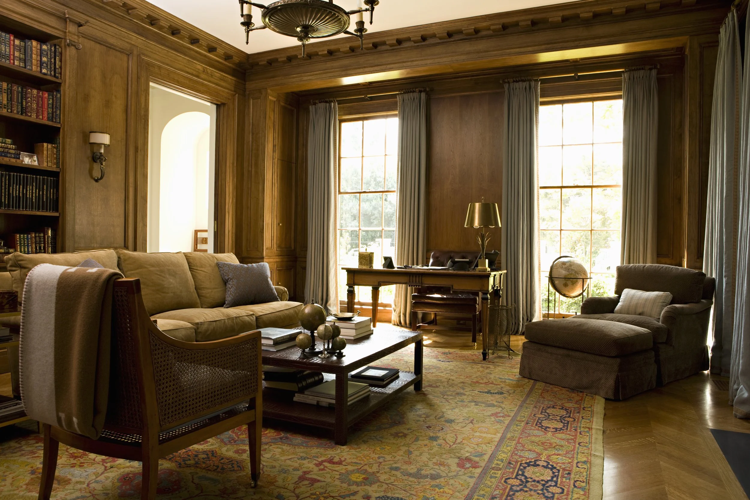 Paneled library with a camel sofa, desk and tall windows with gray draperies