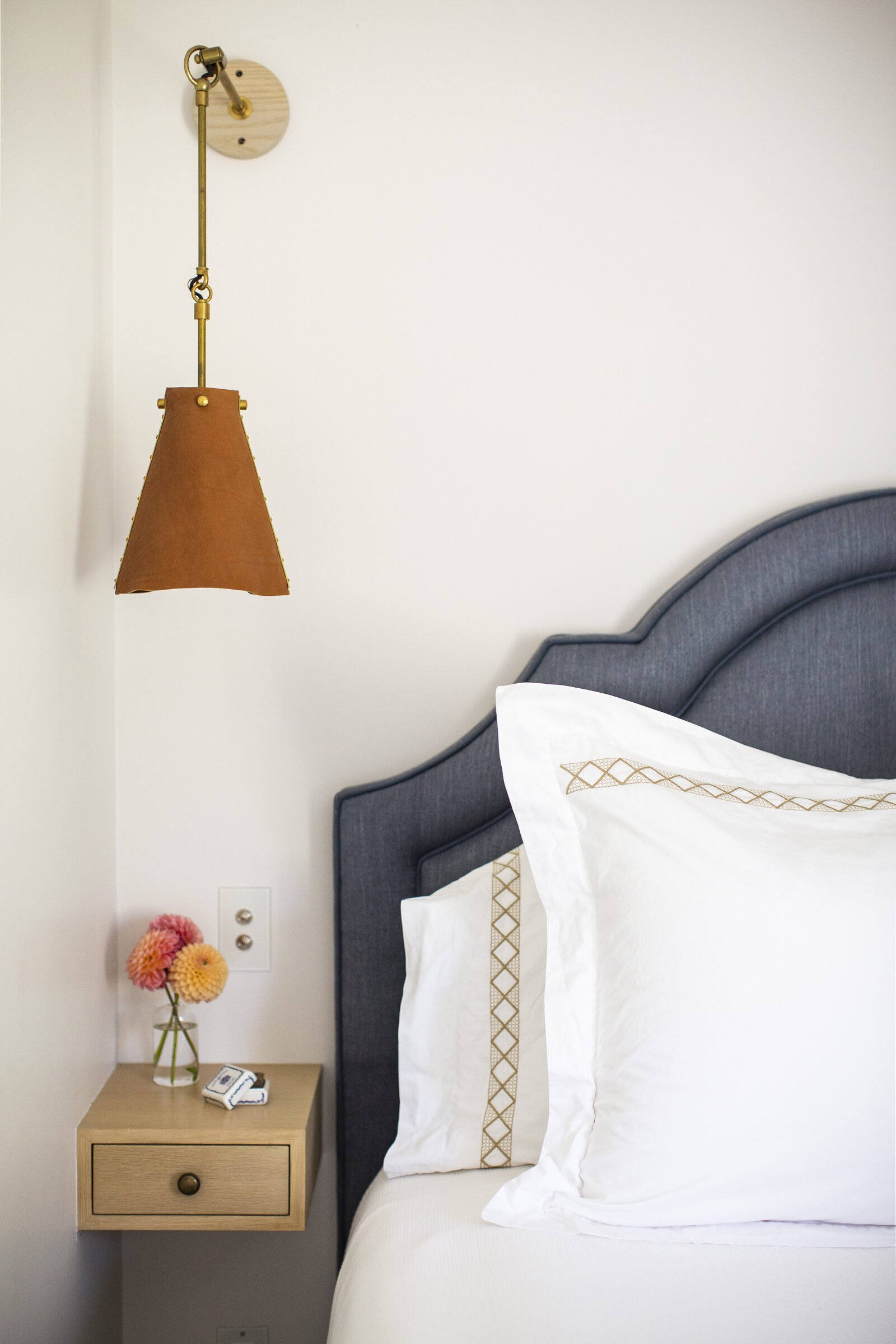 Bed with a navy upholstered headboard and white linens with tan trim, a wood floating nightstand with dahlias and a brass and leather wall light