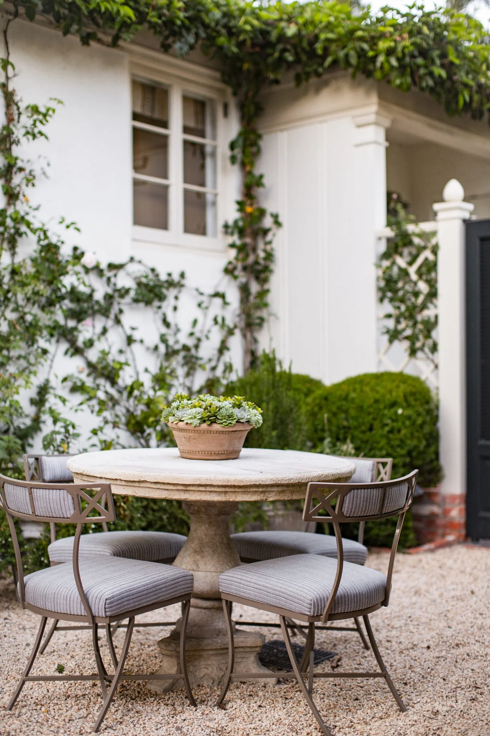 Garden corner with a round stone table, four metal chairs with striped cushions, a potted succulent, clipped boxwood and a vine-covered white wall