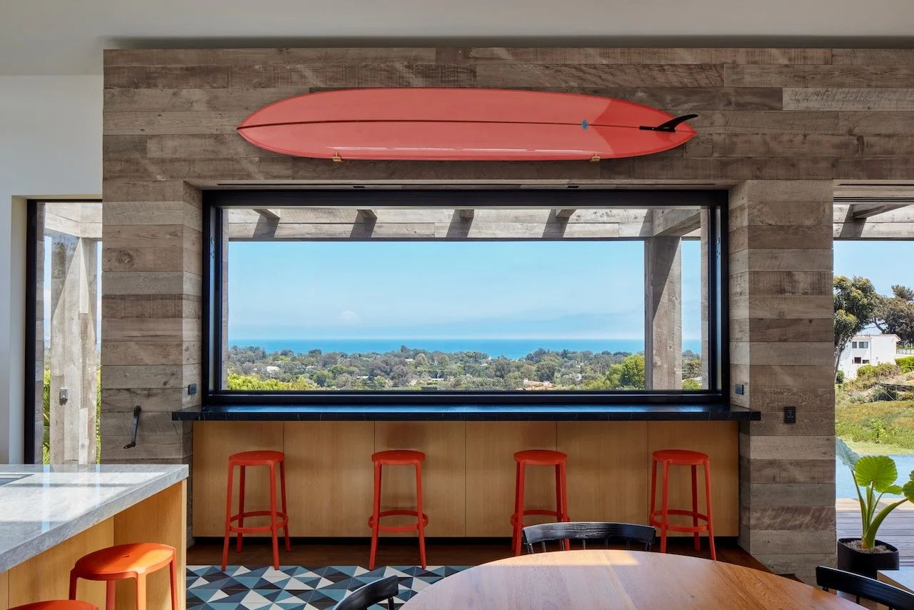 Kitchen counter with red stools under a picture window with an ocean view and a red surfboard on the wall