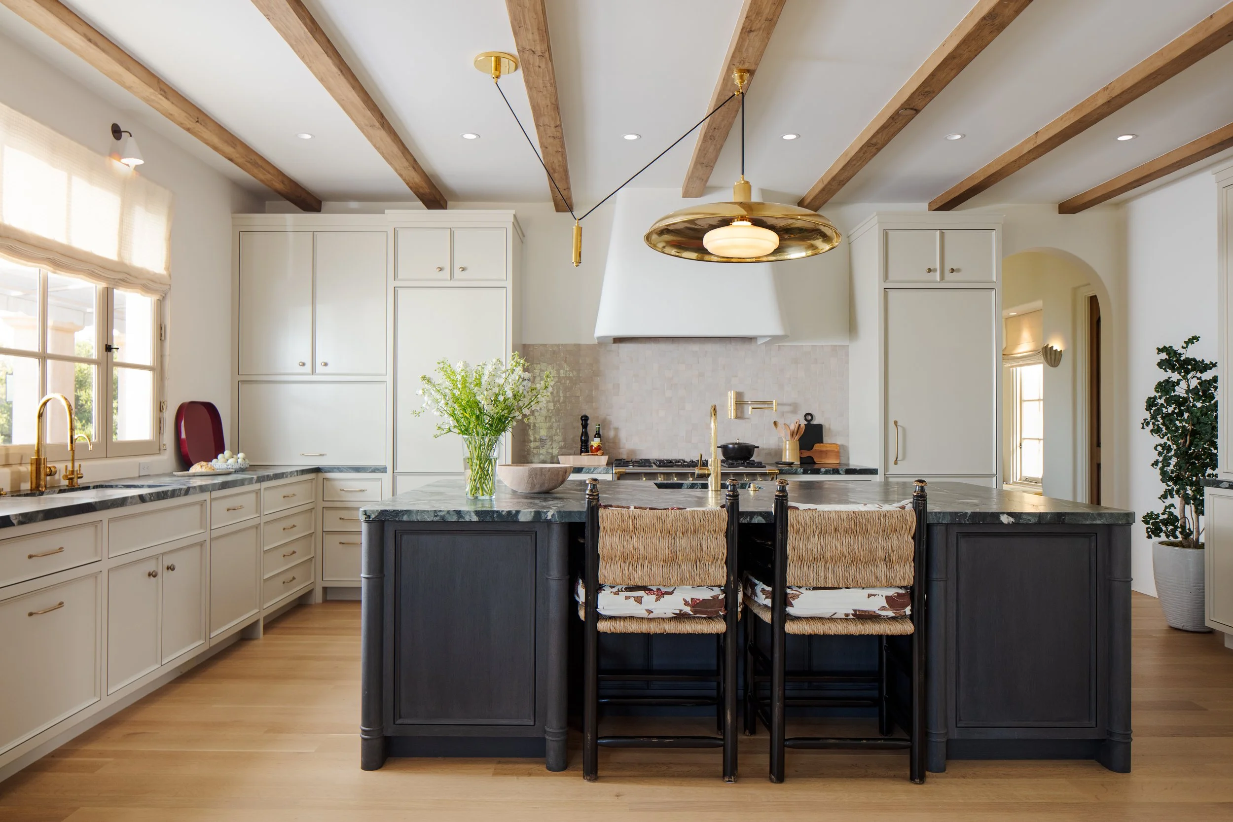 Kitchen with exposed wood beams, white cabinetry, a black island with rush counter stools and a brass pendant