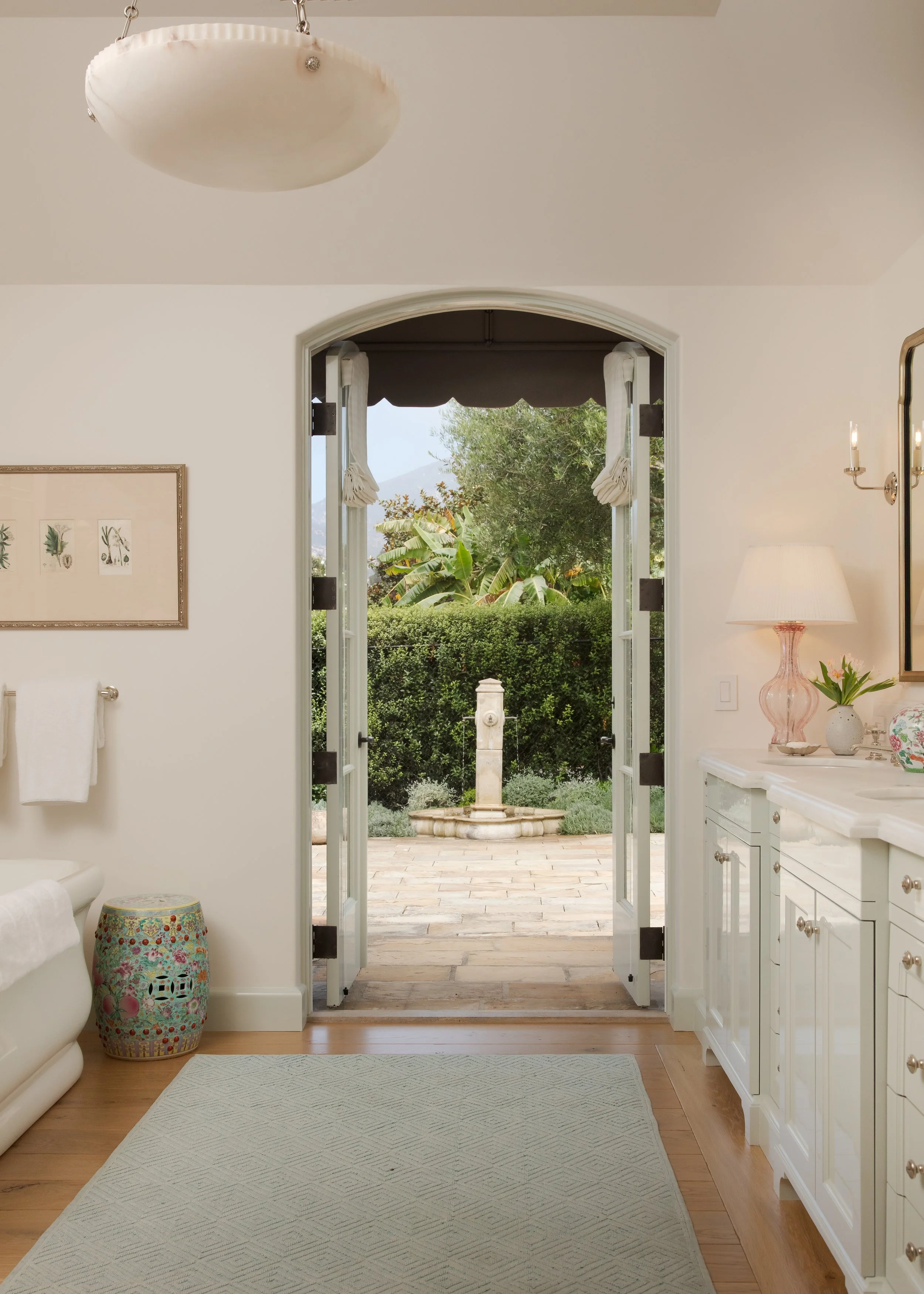 Arched French doors with a scalloped awning open to a garden courtyard with a stone statue, beside a vanity with a pink lamp
