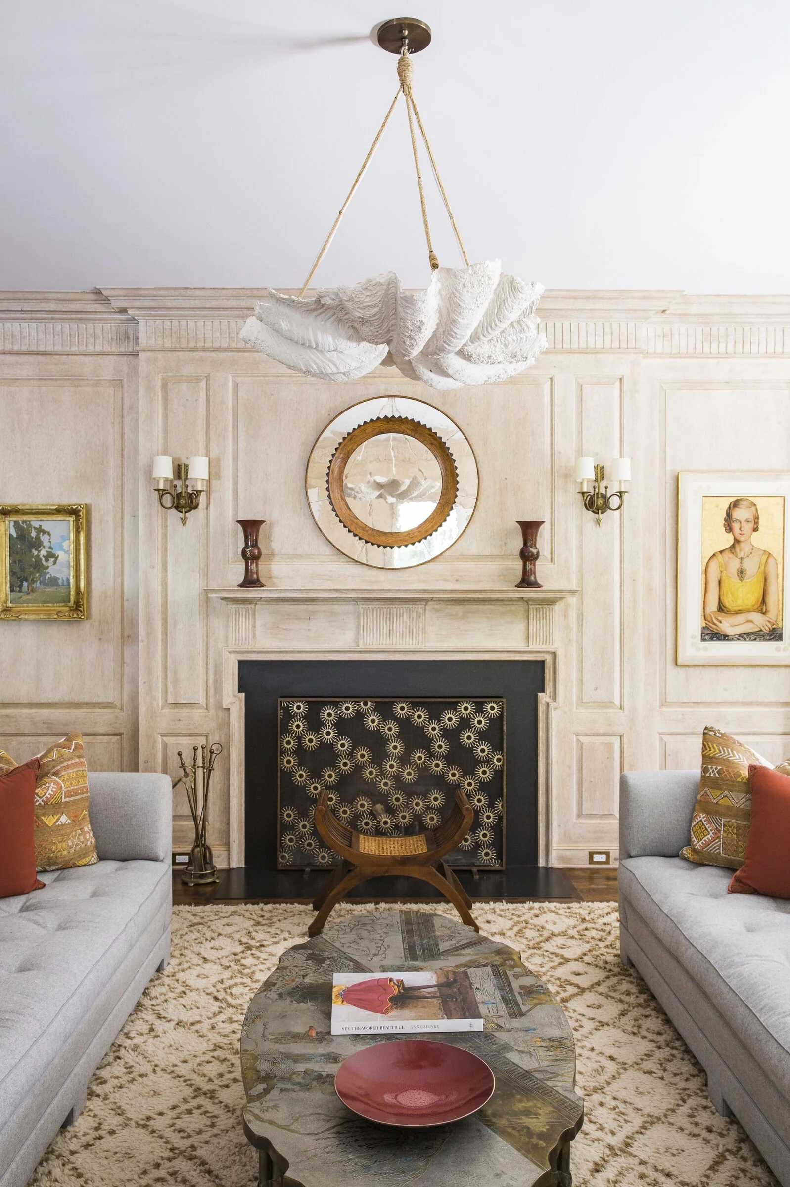 Sitting room with a limed wood paneled fireplace wall, a plaster shell chandelier, two gray tufted sofas, a round mirror and a patterned stone coffee table