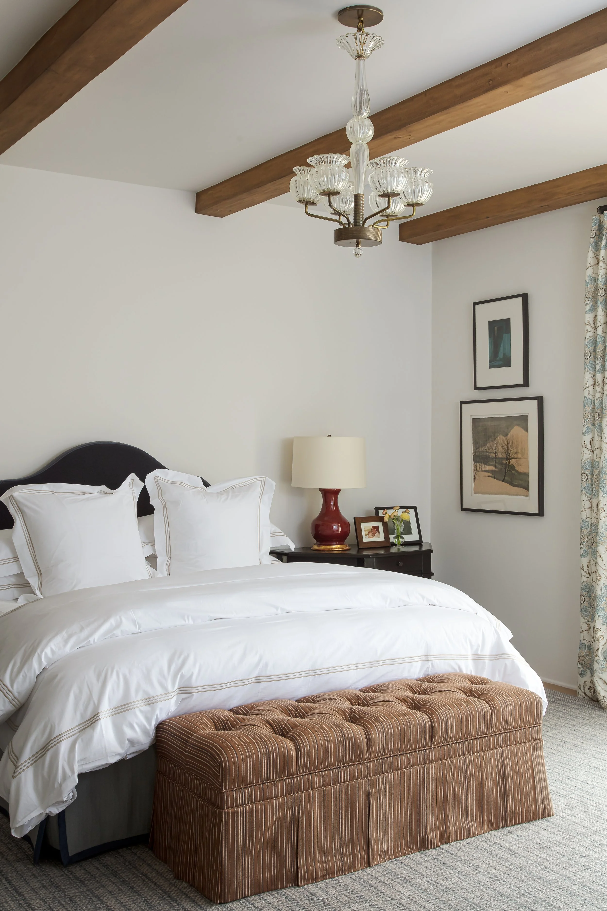Bedroom with a black upholstered headboard, white bedding, a red ceramic lamp and framed art