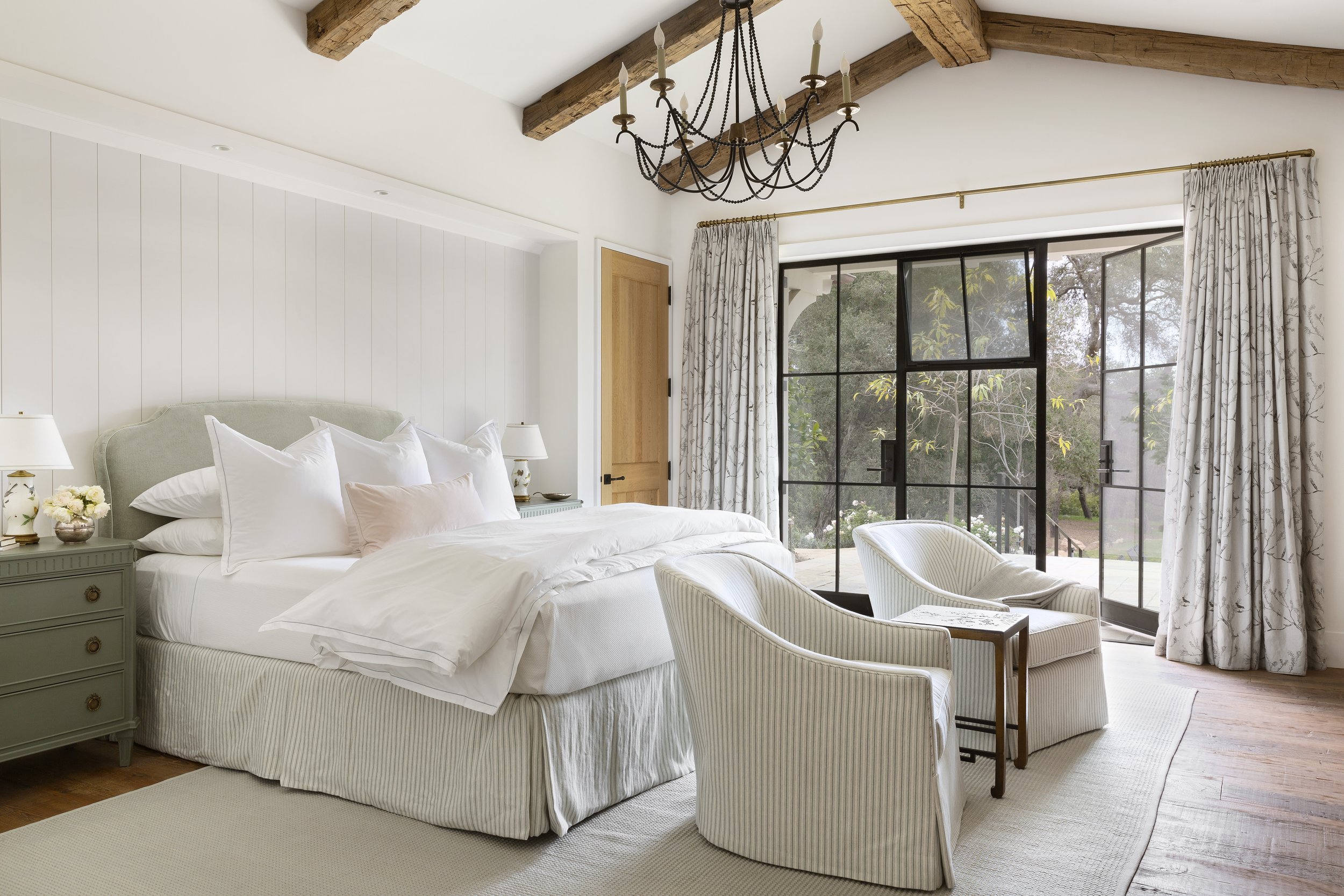 Primary bedroom with exposed beams, steel-framed doors, striped armchairs and floral curtains