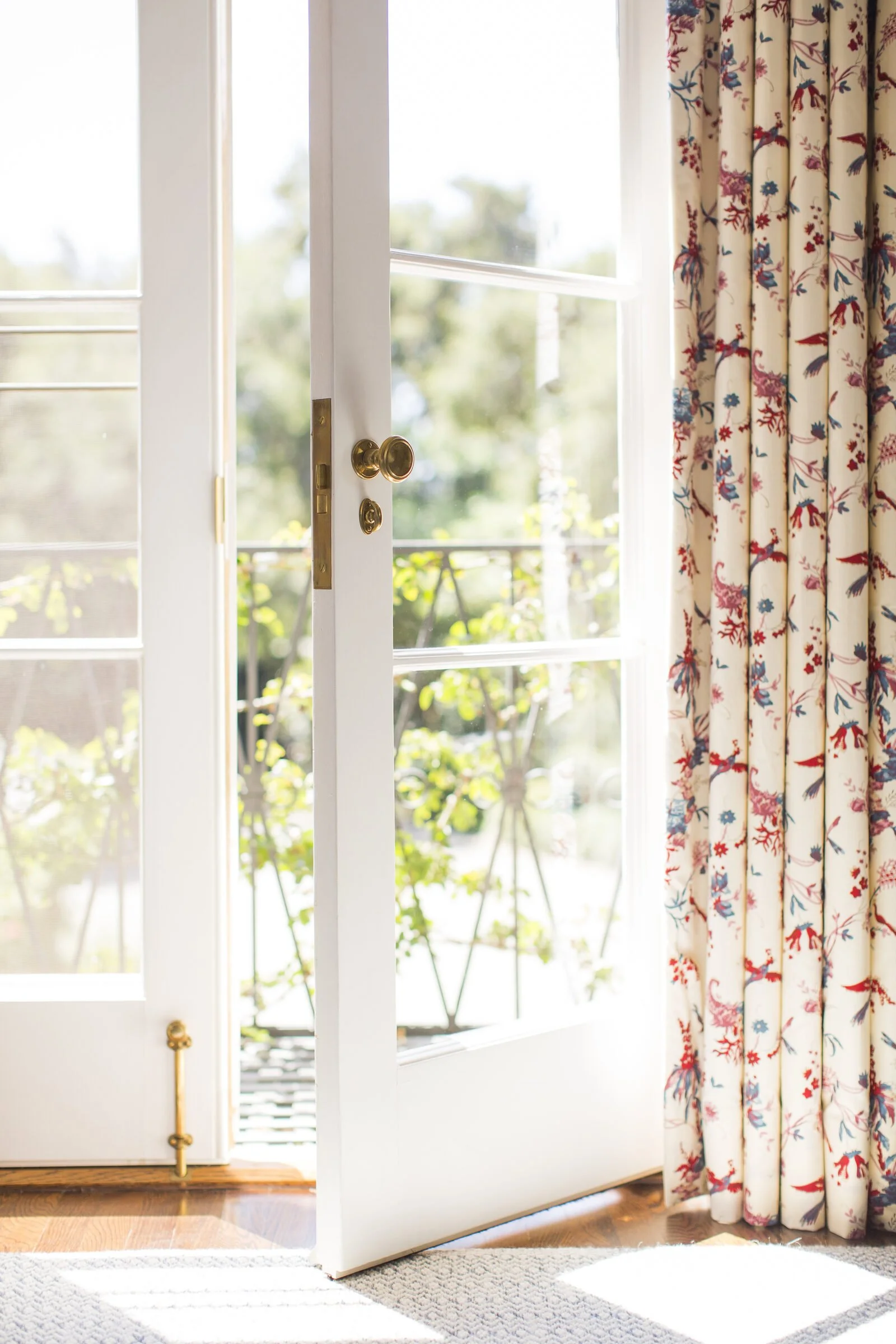 Open French door with a brass knob and a long red-and-blue floral curtain looking out to a garden