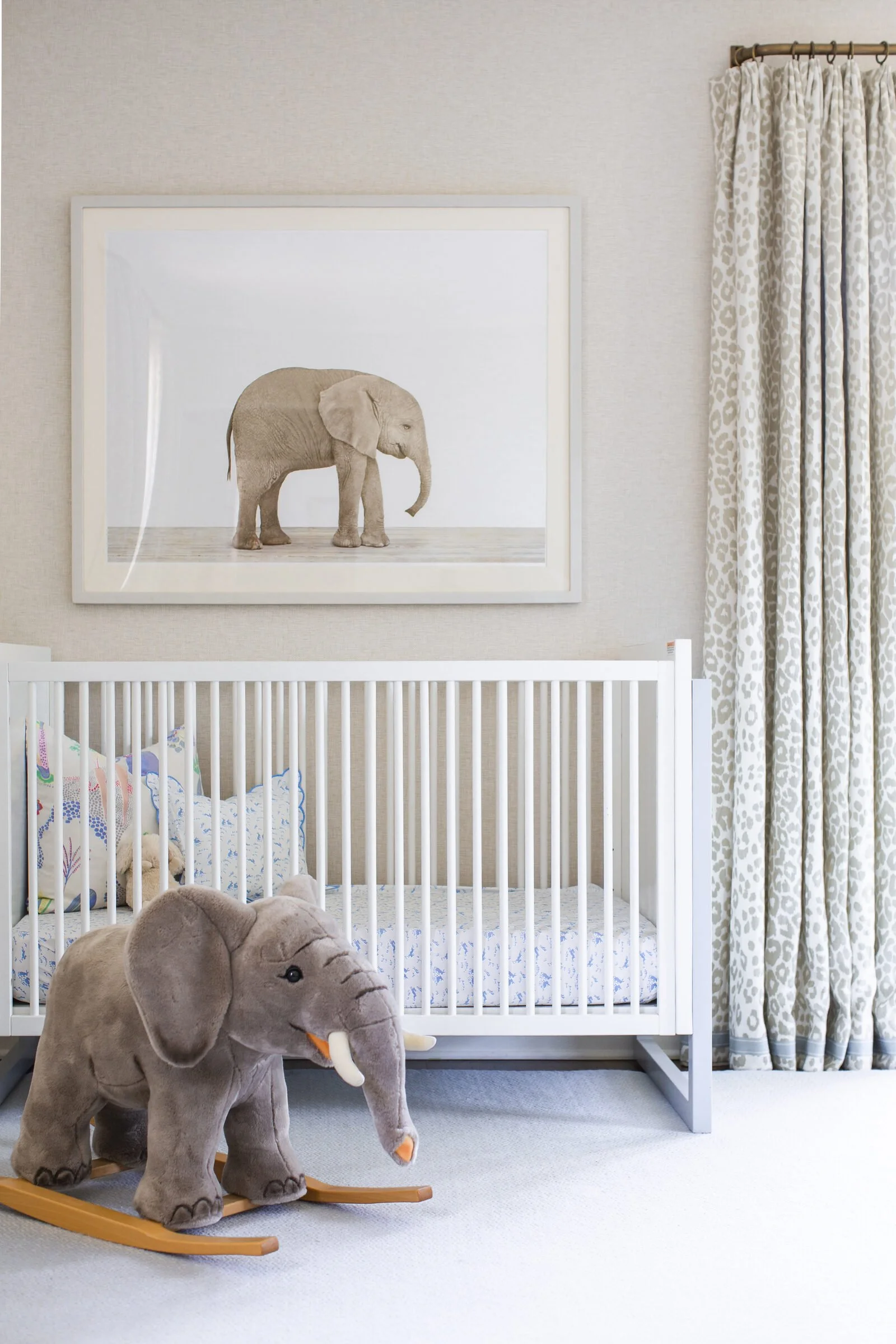 Nursery crib with a framed elephant photograph, a plush elephant rocker and gray leopard-print curtains