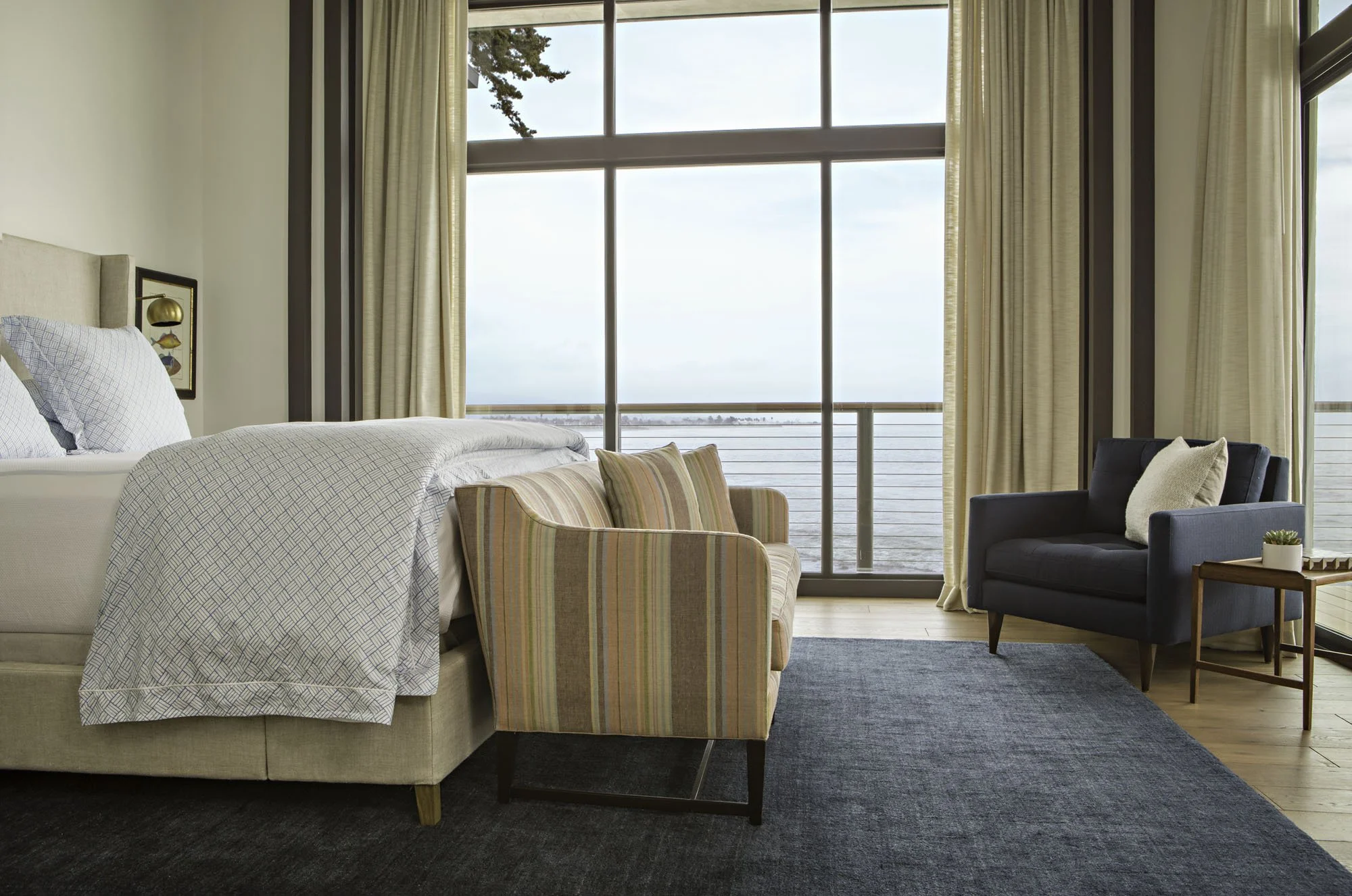 Bedroom with a striped daybed, a navy armchair, a navy rug and tall windows looking out to the sea