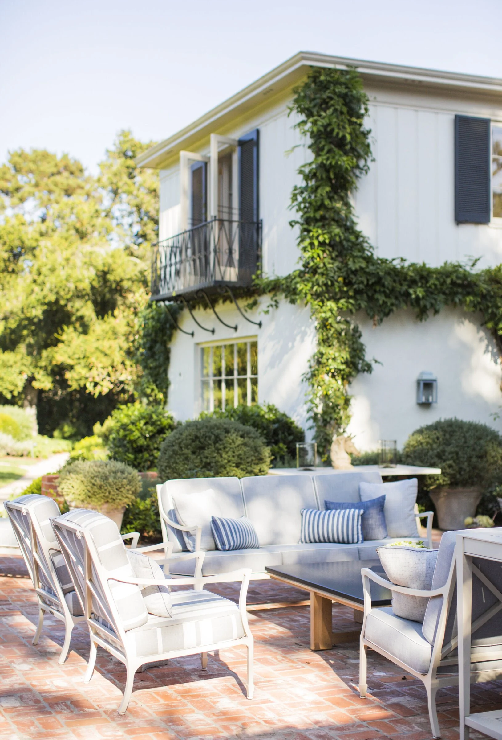 Brick patio beside a vine-covered white house with a wrought iron balcony, a striped outdoor sofa, white armchairs and a low table