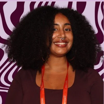 A woman with curly hair and a nose piercing smiling in front of a purple and white patterned background.