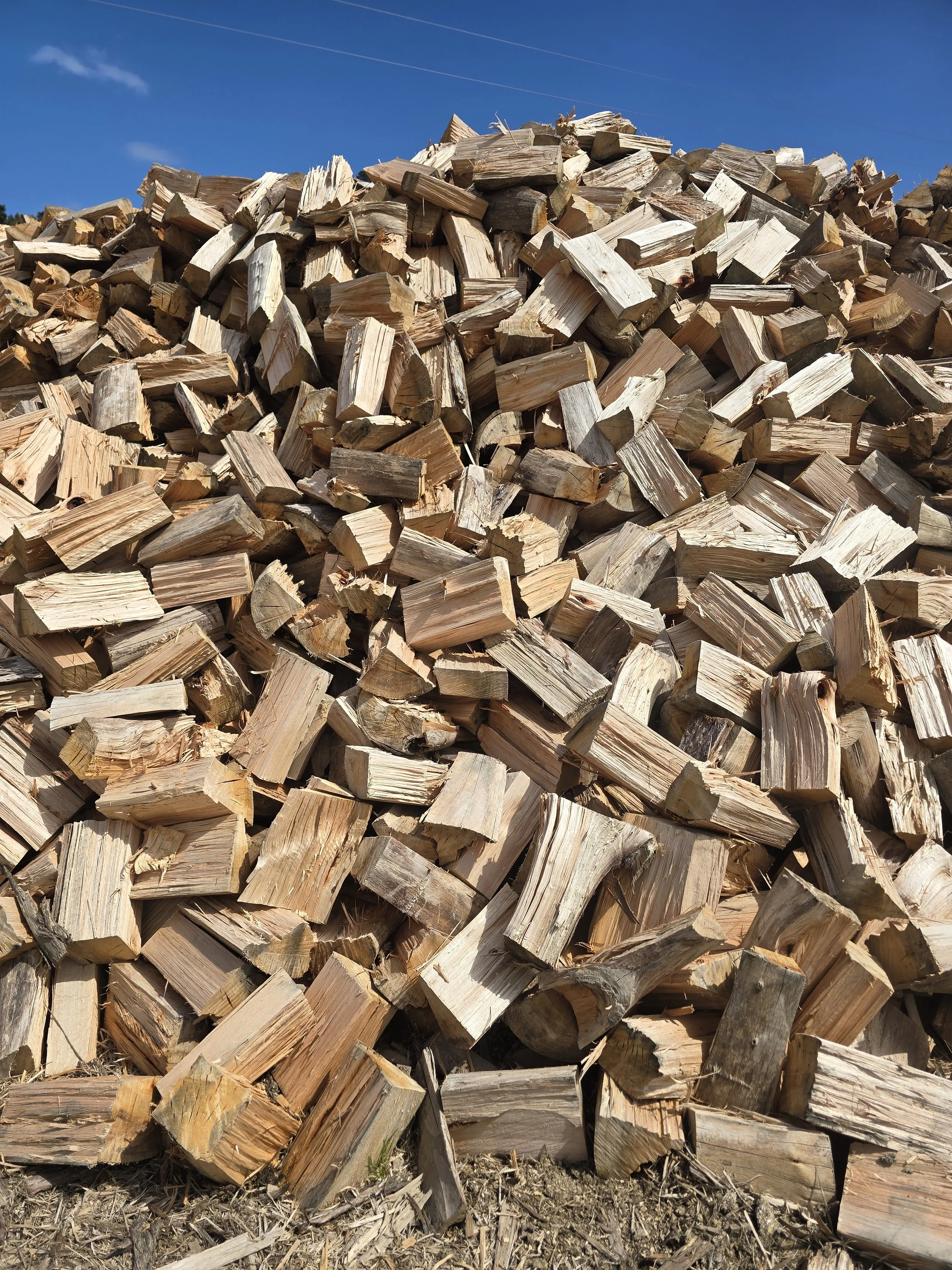 A large pile of chopped firewood stacked outdoors under a clear blue sky.