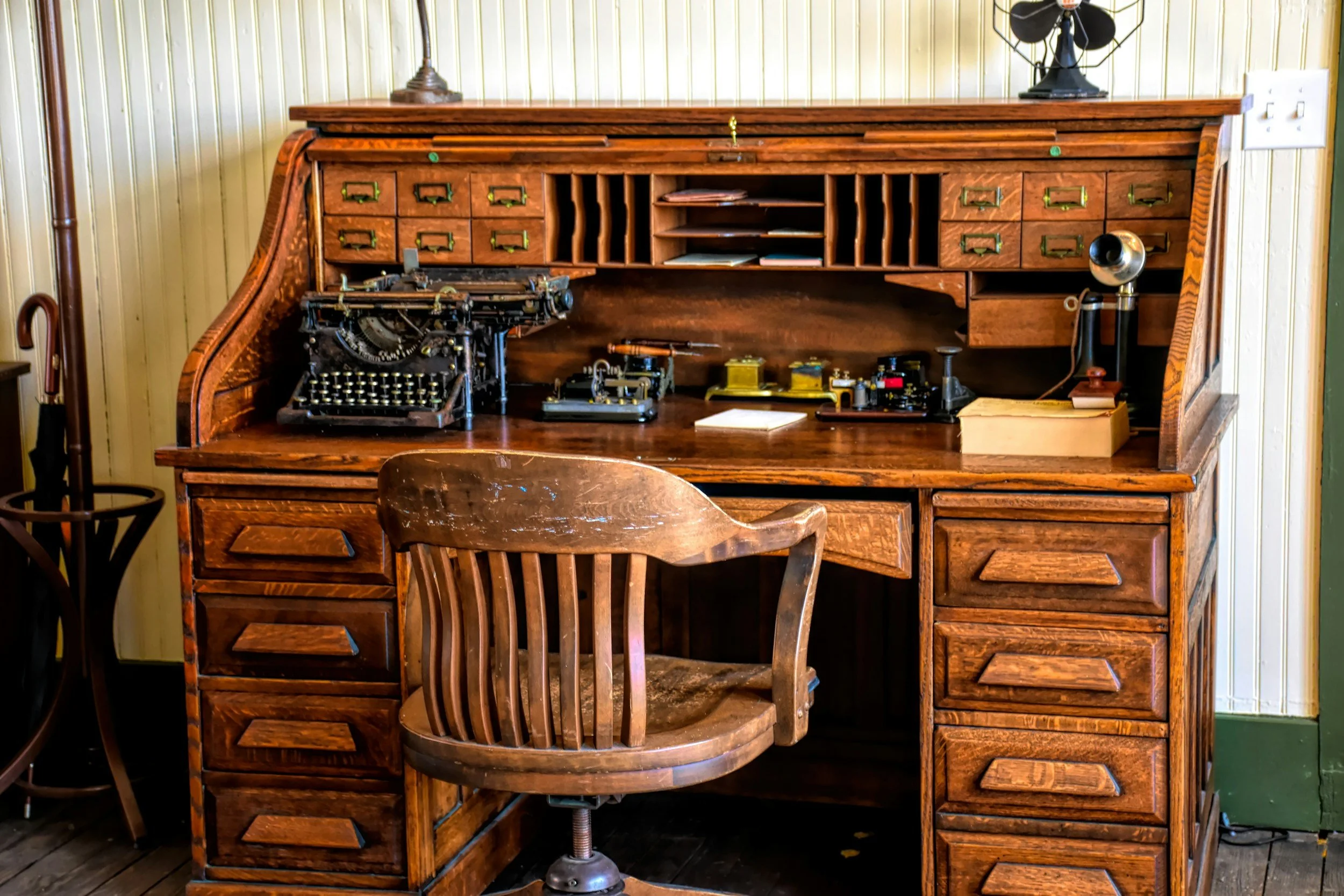Antique wooden rolltop writing desk with vintage typewriters, stationery compartments, and a wooden chair.