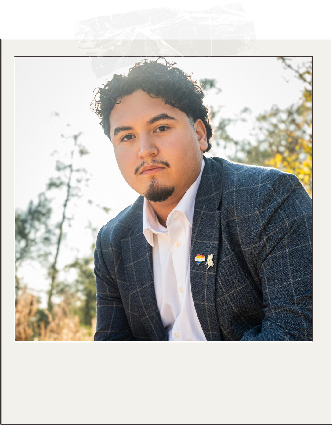 A young man with curly dark hair, light skin, and a small goatee, dressed in a white shirt and a dark plaid blazer, outdoors during daytime with trees in the background, wearing rainbow pride and protest pins.