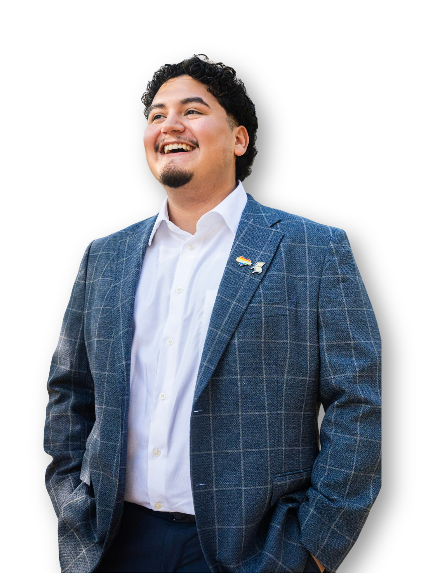A man wearing a blue checkered suit, white shirt, and a rainbow pride pin smiling with his hands in his pockets.