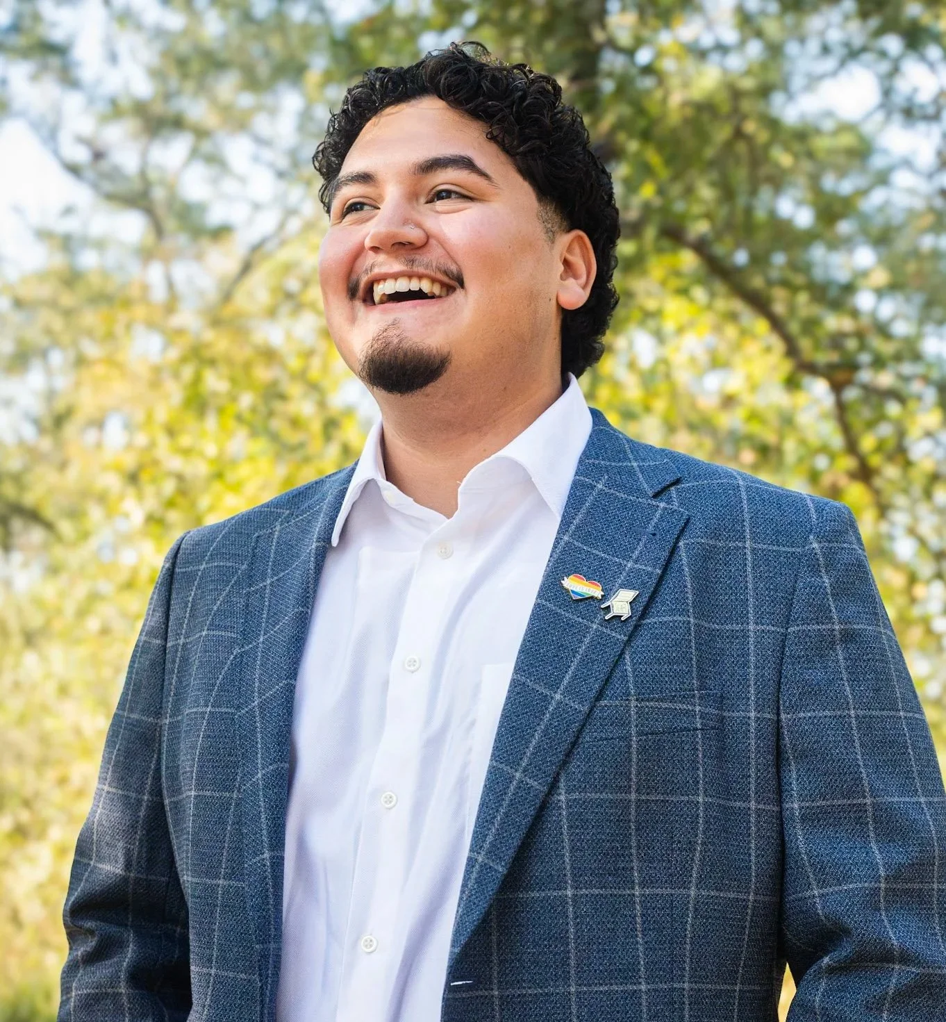 A smiling man with dark, curly hair wearing a white dress shirt and a blue checkered blazer, standing outdoors with green trees in the background.
