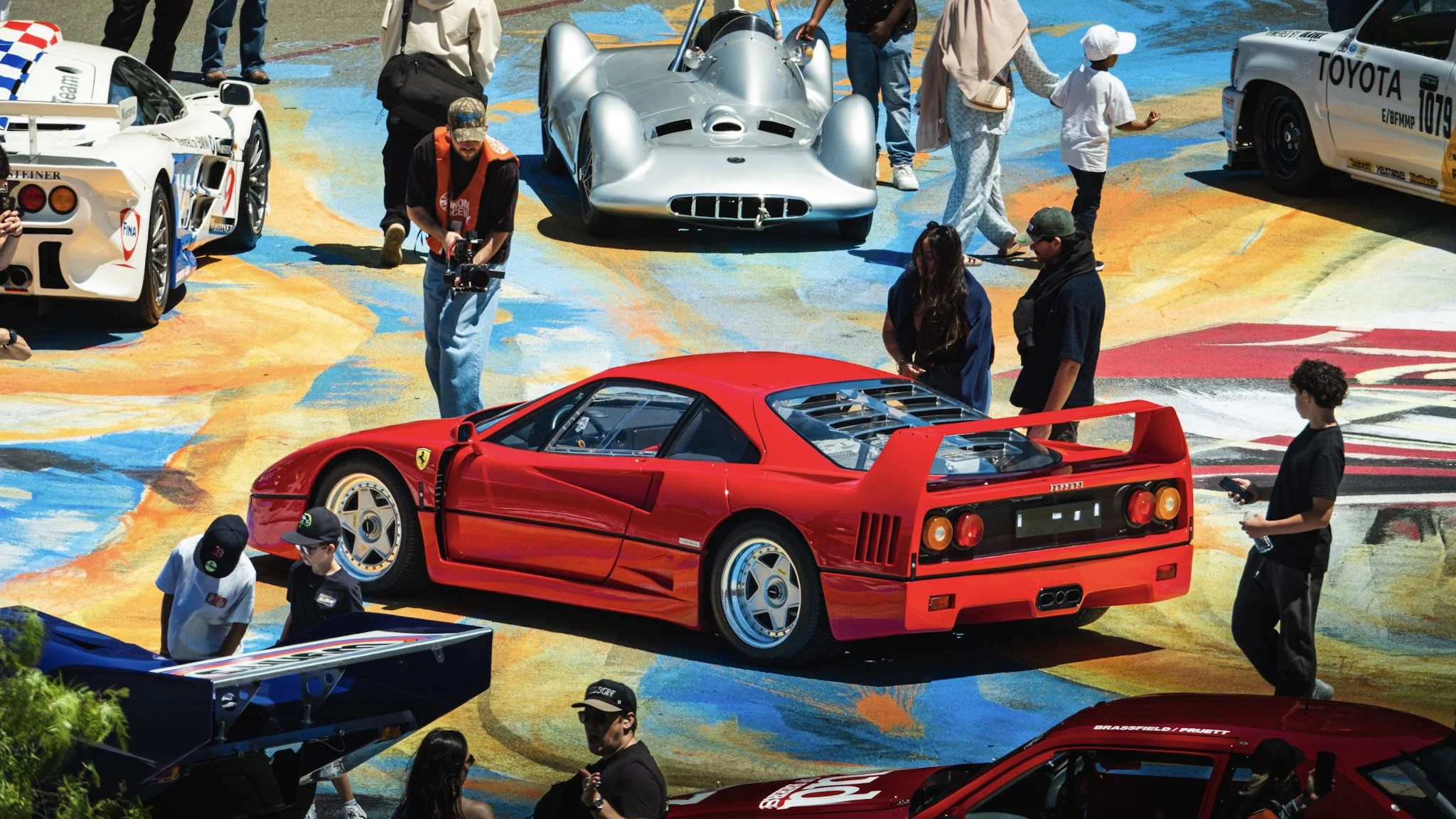 Various classic and exotic cars, including a red Ferrari F40 and a silver classic race car, are parked and displayed outdoors during a car show. People are walking around, taking photos, and observing the vehicles on a colorful painted ground.