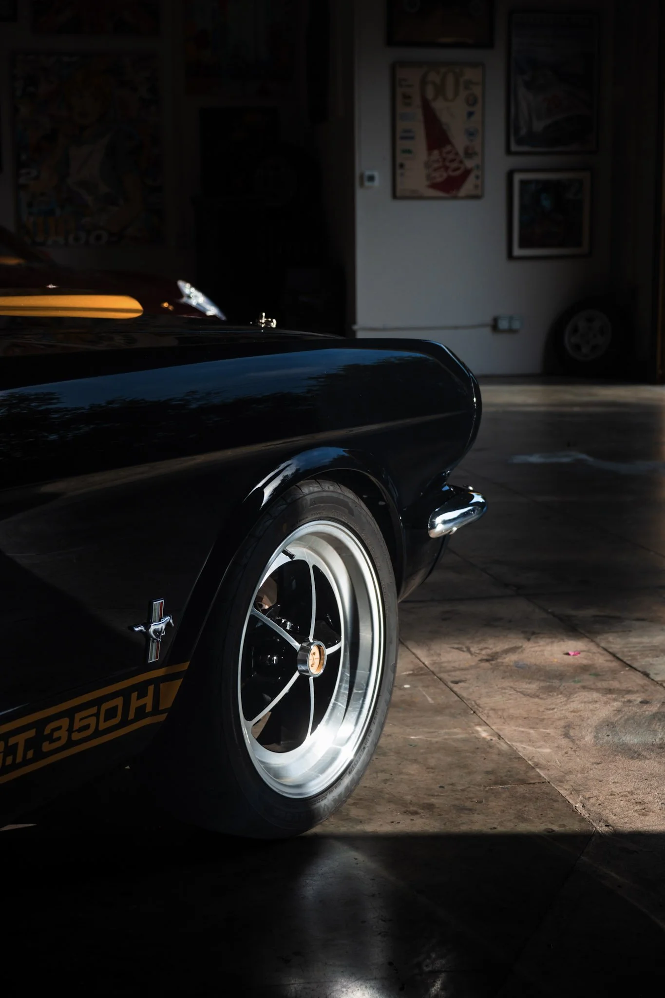 Partial view of a black vintage car with white-rimmed wheel, parked inside a garage. The garage wall has framed posters and artwork, with a tire visible on the floor in the background.