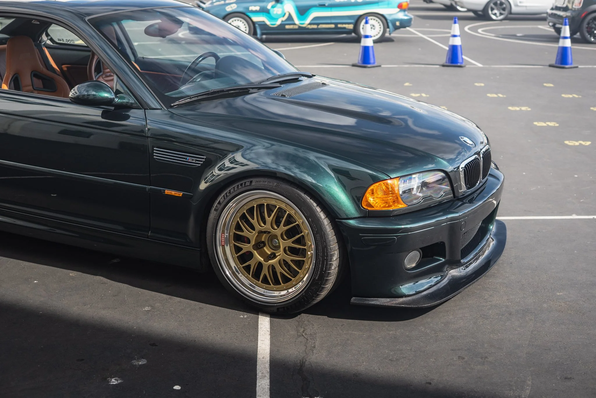 A black BMW M3 sports car with gold wheels parked in a parking lot, with several other cars and blue traffic cones in the background.