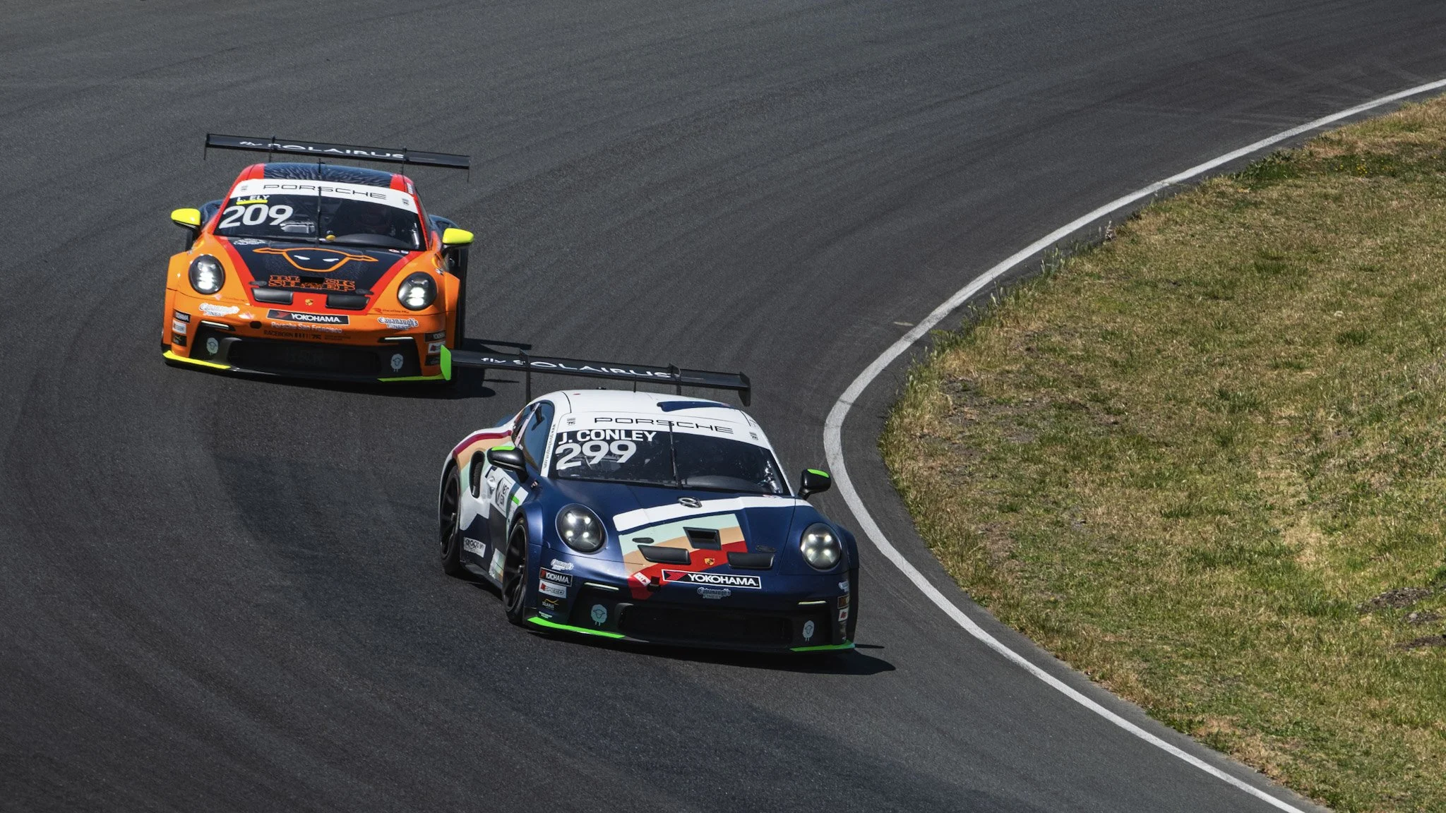 Three race cars navigating a turn on a racetrack, with the leading car being orange and black and the others blue and white, on a curved section of the asphalt track bordered by grass.