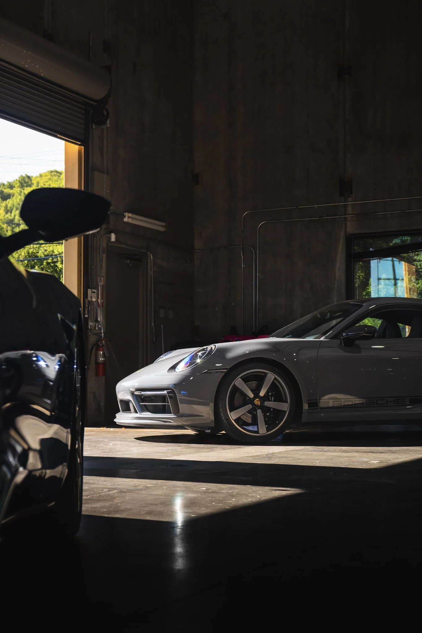 Luxury silver Porsche sports car parked inside a garage with a dark private wall and open garage door showing green trees outside.